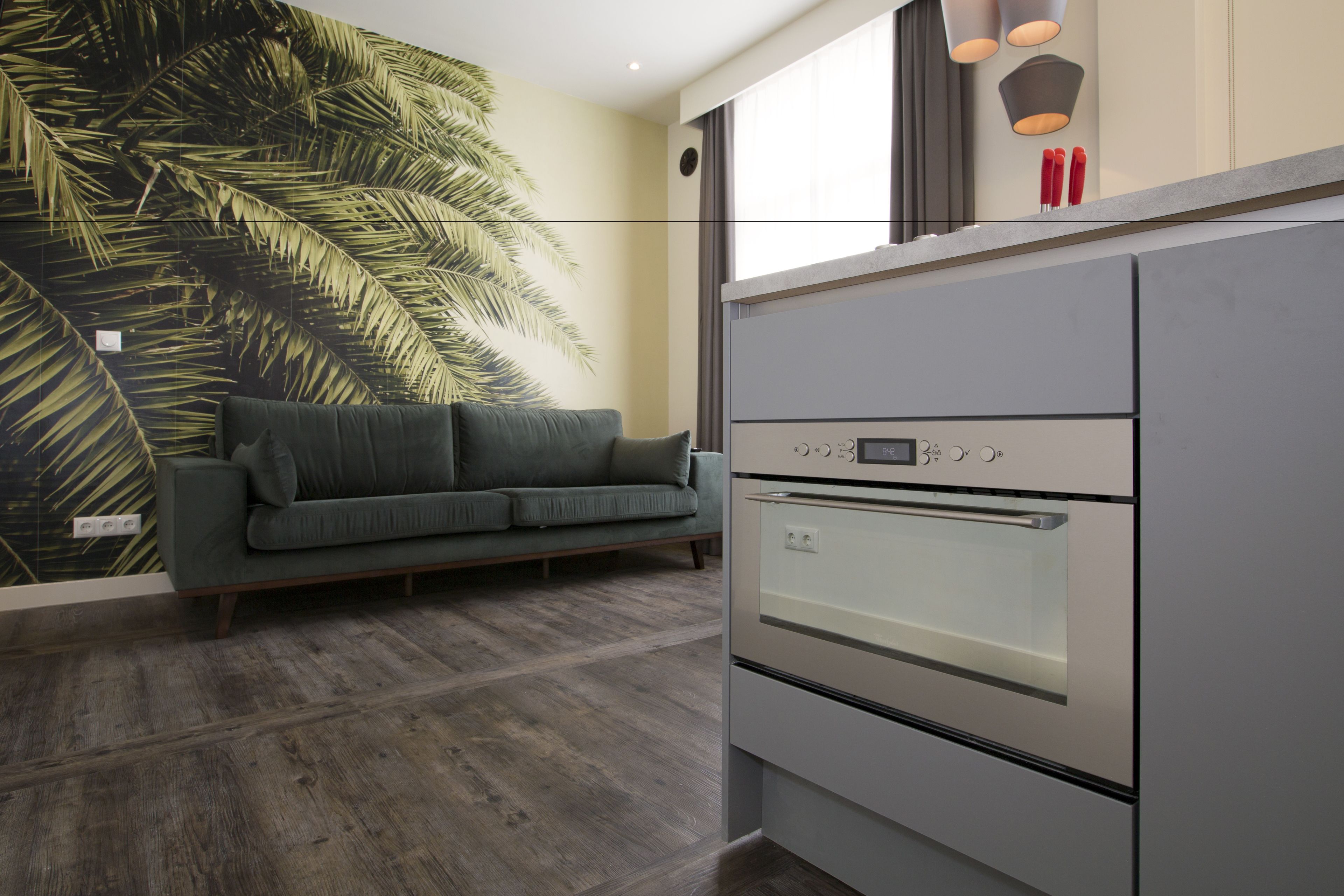 A modern kitchen with a built-in oven, a grey countertop, and a green sofa against a wall with a large leaf pattern.