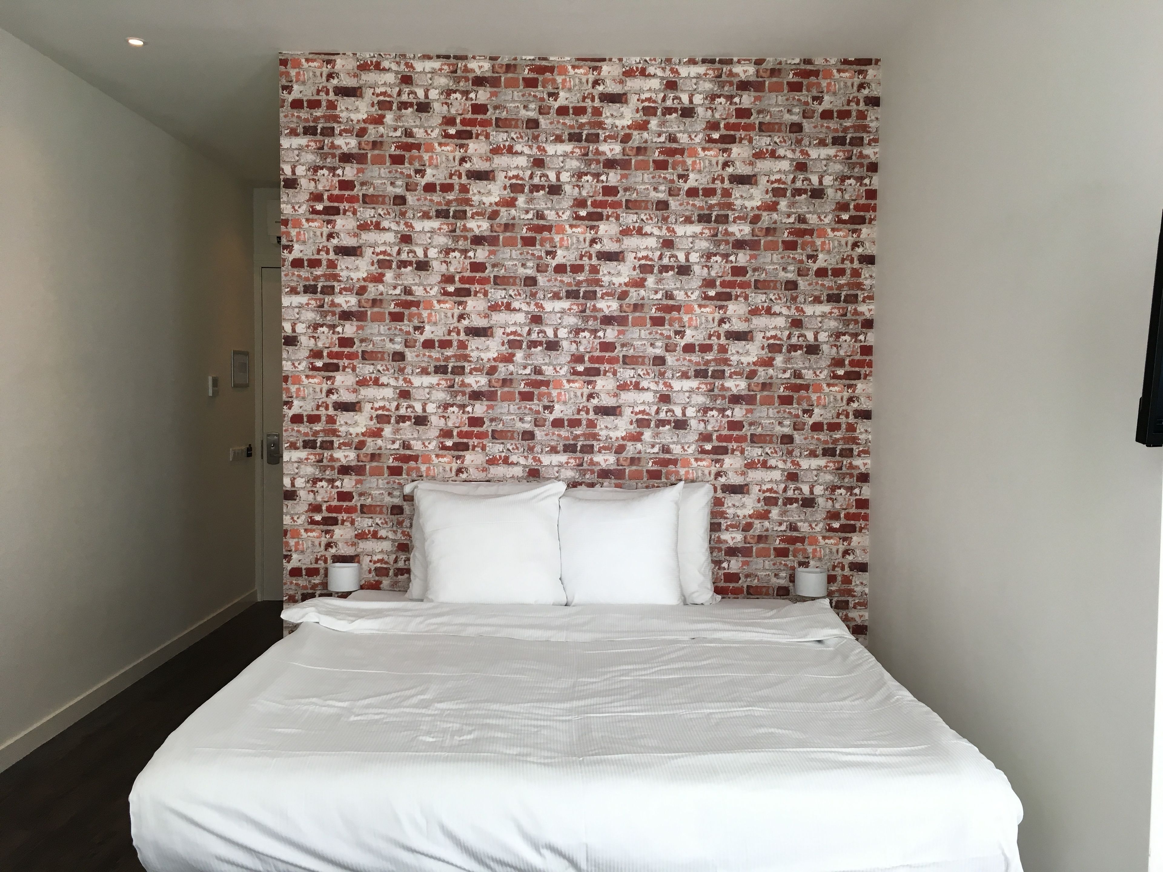 Een hotelkamer met een hoofdeinde met een bakstenen patroon, een groot bed met wit beddengoed, en twee nachtkastjes met lampen.
