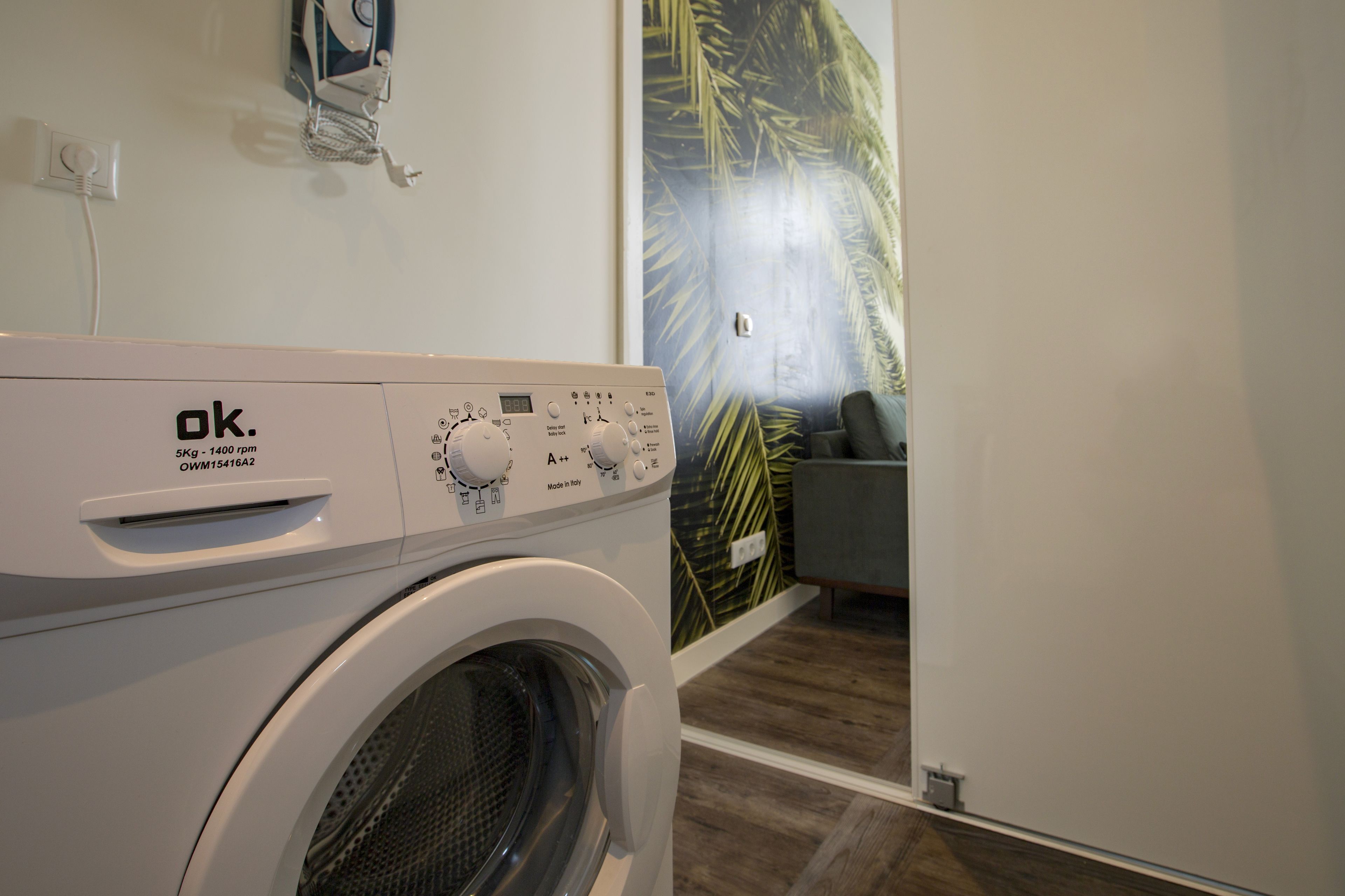 A front-loading washing machine in a room with a wooden floor and a large window with a view of palm trees.