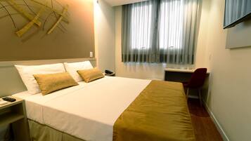Una cama bien hecha con ropa de cama blanca y dorada, una mesita de noche con un teléfono y una silla roja en una habitación de hotel con un ventanal grande y un televisor en la pared.