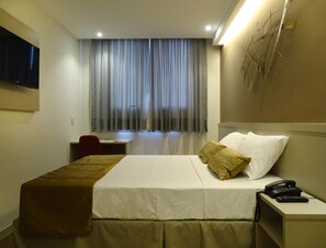 Habitación de hotel con cama, mesita de noche con teléfono, escritorio con silla, televisor montado en la pared y ventana con cortinas.