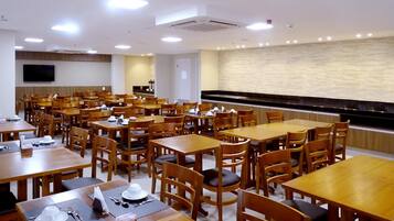 A dining area with multiple wooden tables and chairs, a flat-screen TV mounted on the wall, and a stone-textured wall in the background.
