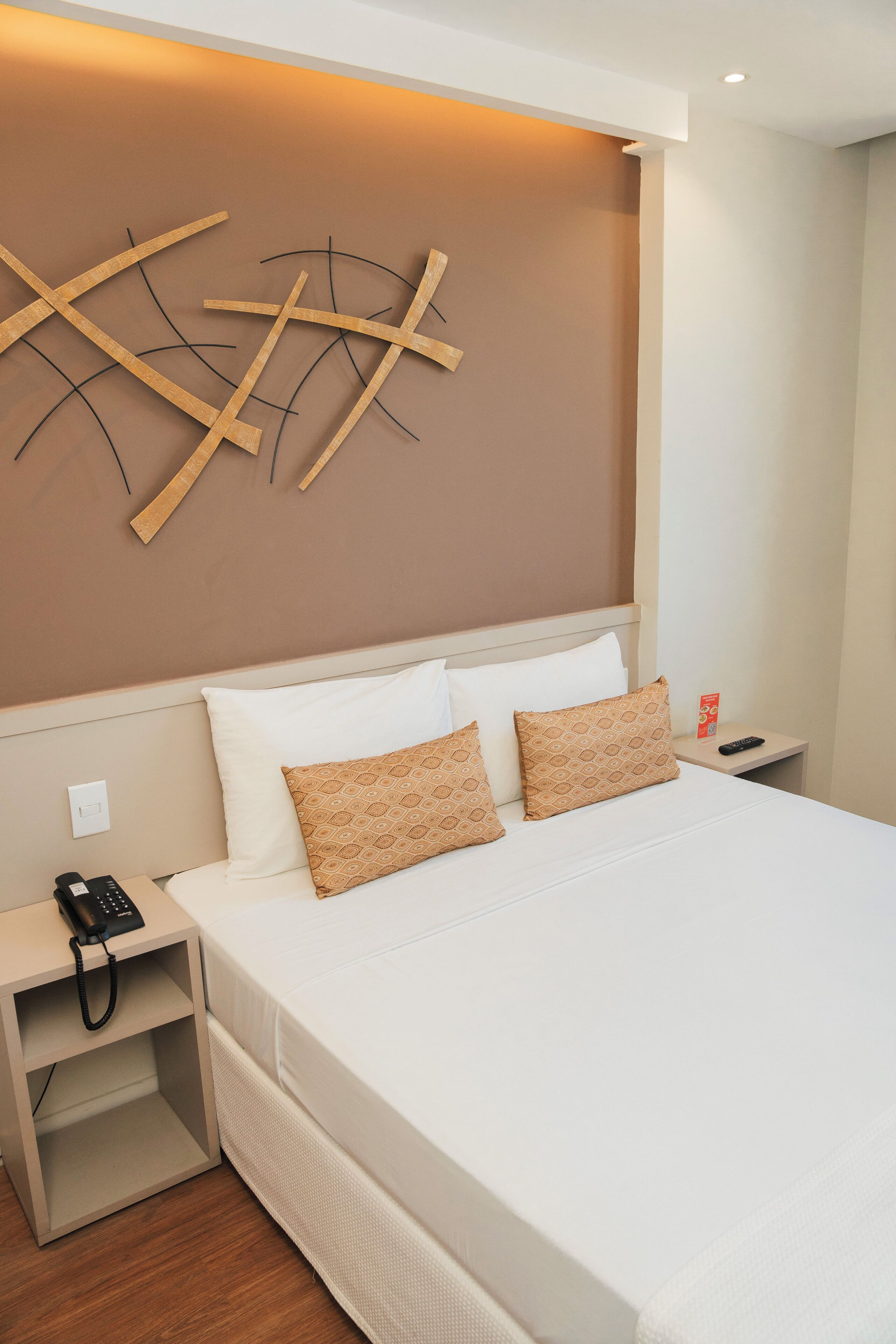 Quarto de hotel com uma cama grande, mesa de cabeceira com telefone e luminária fixada na parede.