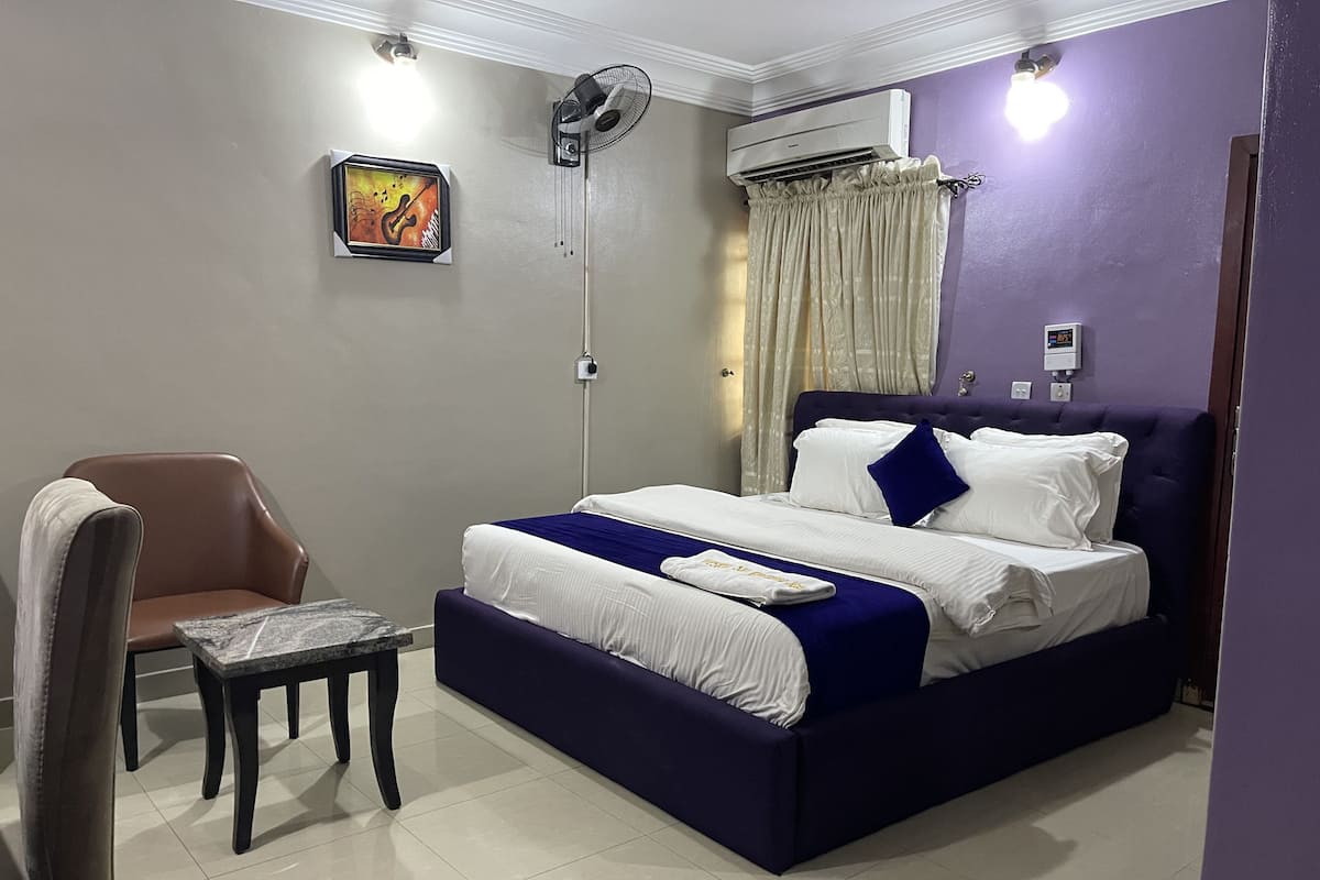 A neatly arranged bedroom with a bed, a chair, a small table, an air conditioner, and a wall-mounted artwork.