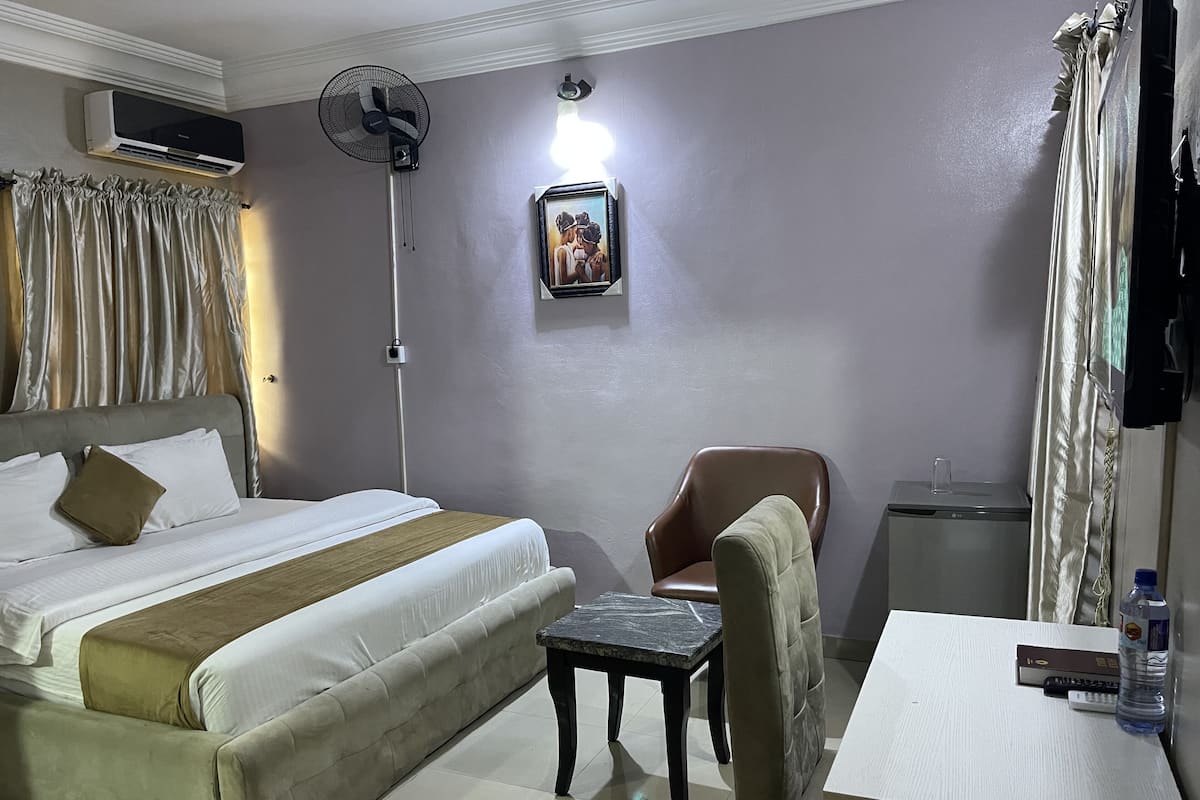 A hotel room with a bed, a chair, a small table, an air conditioner, a standing fan, and a picture on the wall.