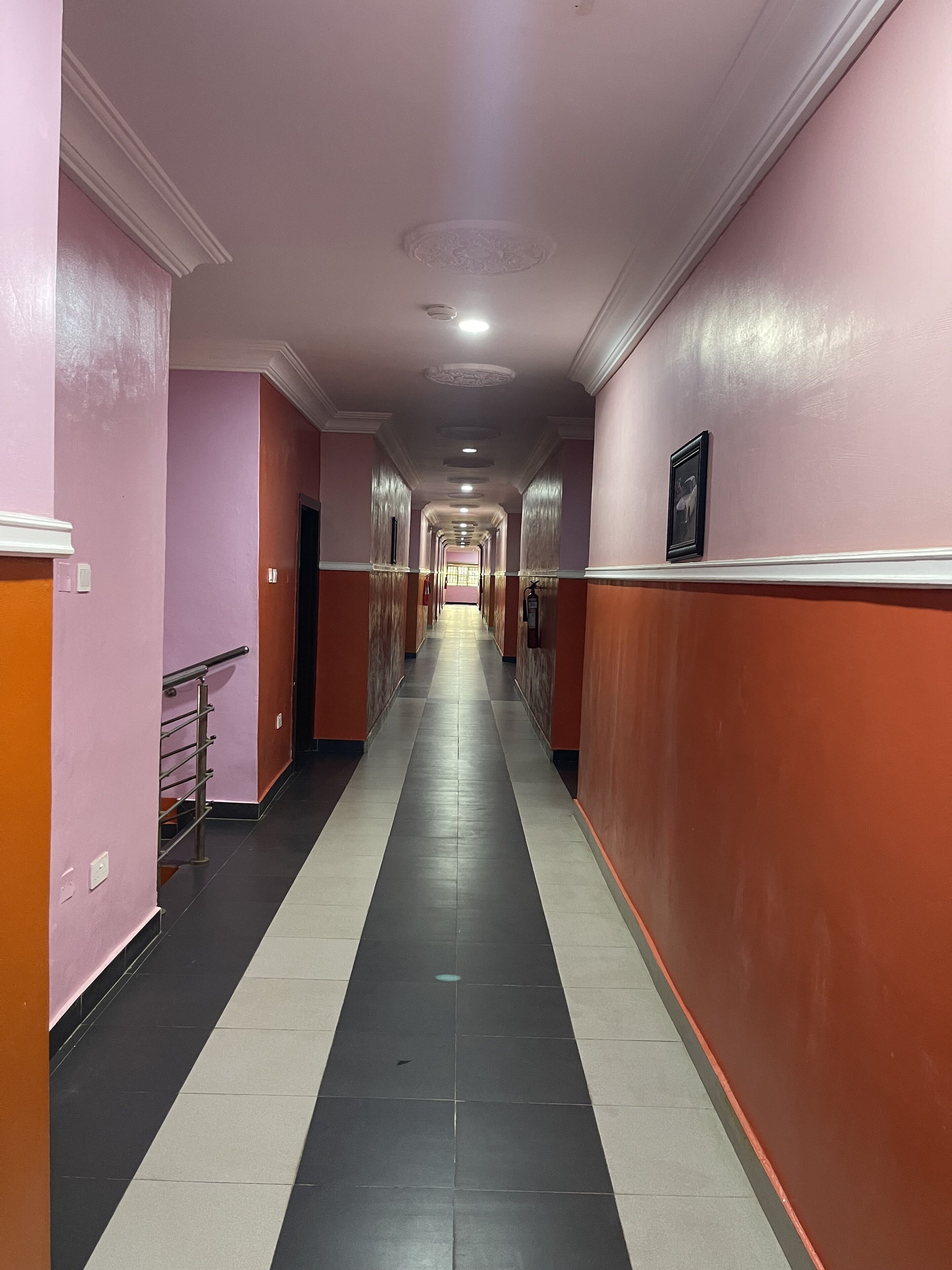 A long, narrow hallway with alternating pink and orange walls, a tiled floor, and a handrail on the left side.