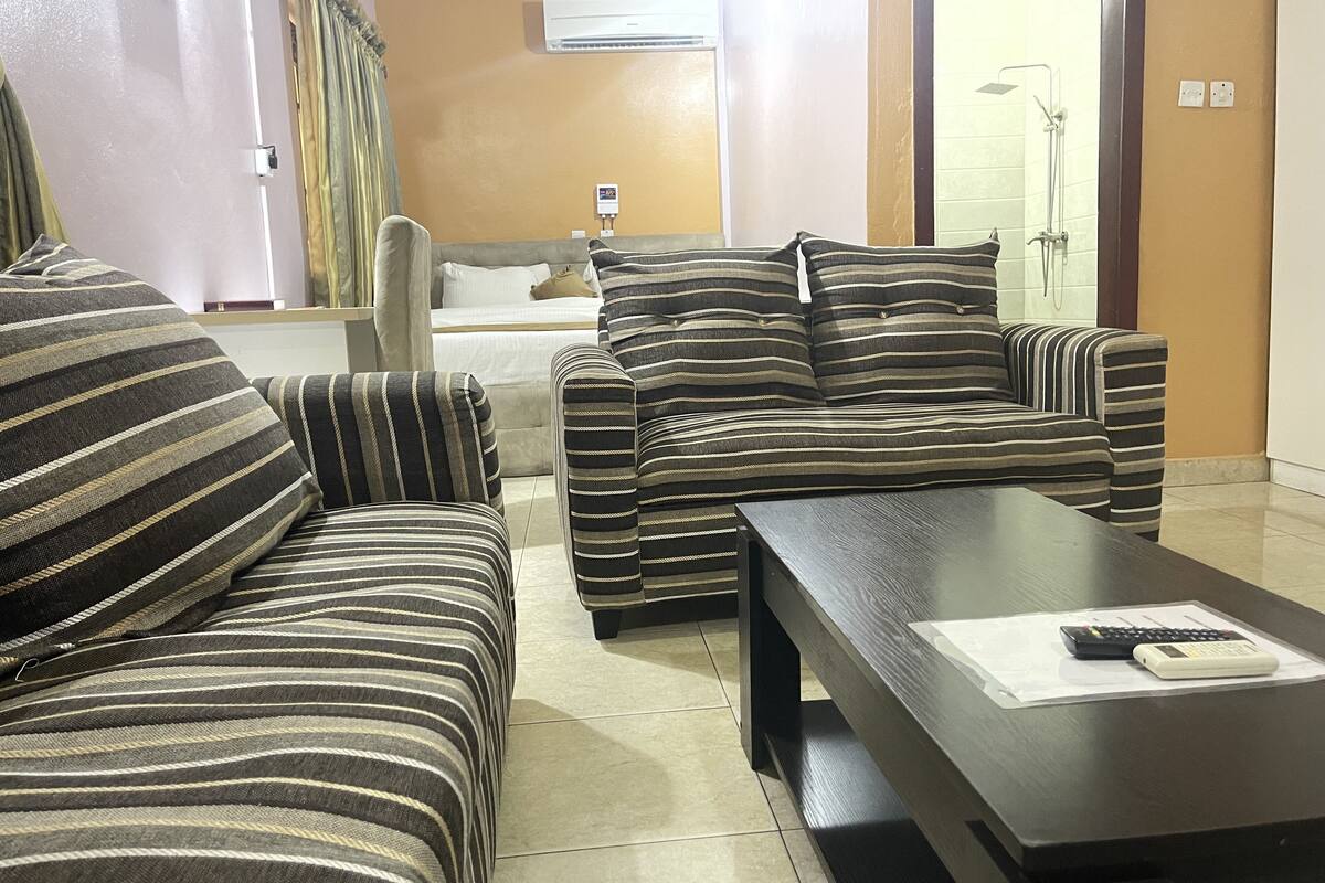 A living room with striped sofas, a dark wooden coffee table, and a bed with white bedding in the background.