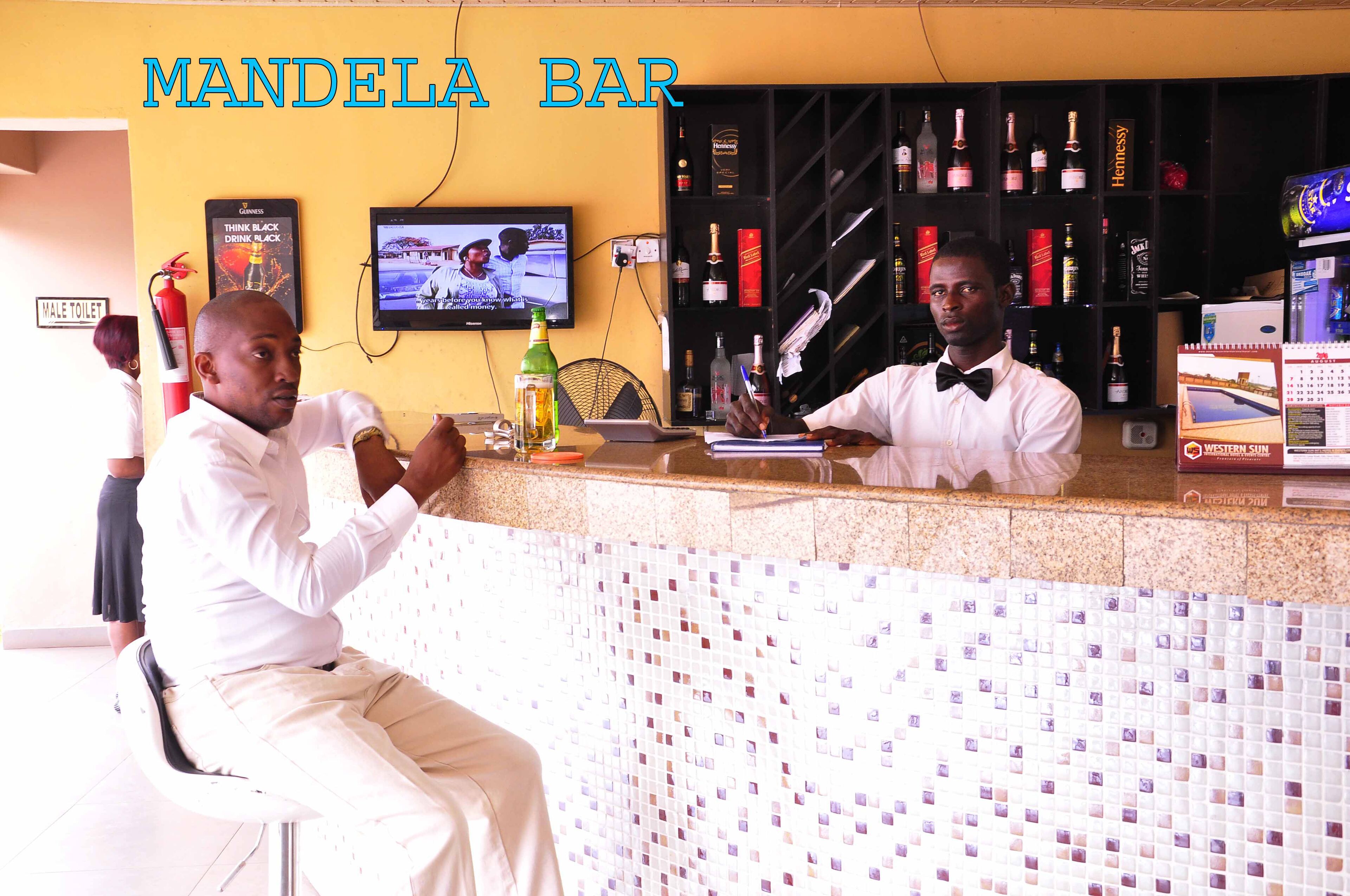 Un bar llamado 'Mandela Bar' con una pared de azulejos, un televisor y estantes llenos de botellas de licores.