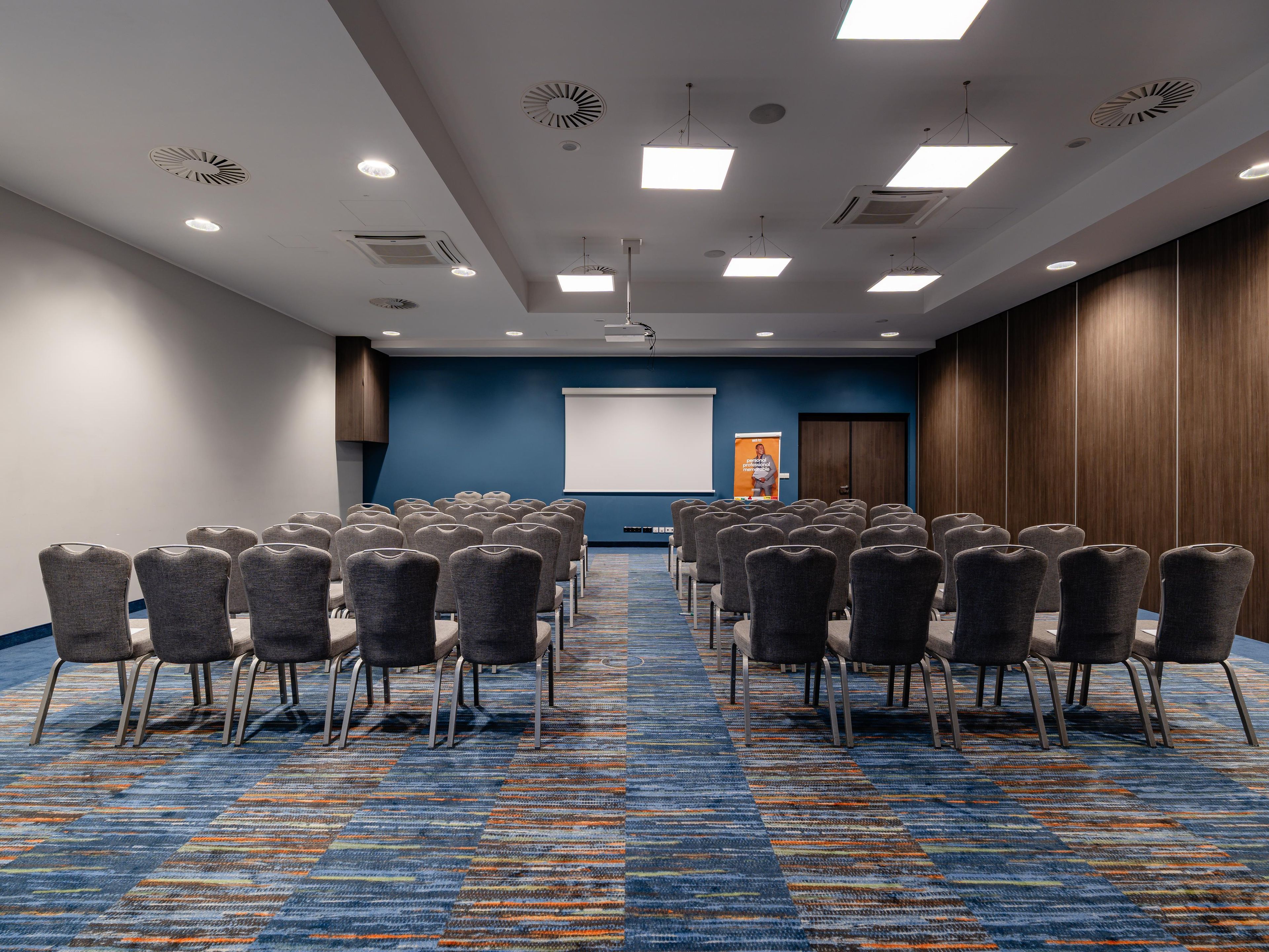 Uma sala de conferências com fileiras de cadeiras, uma tela de projetor e um ecrã de apresentação.