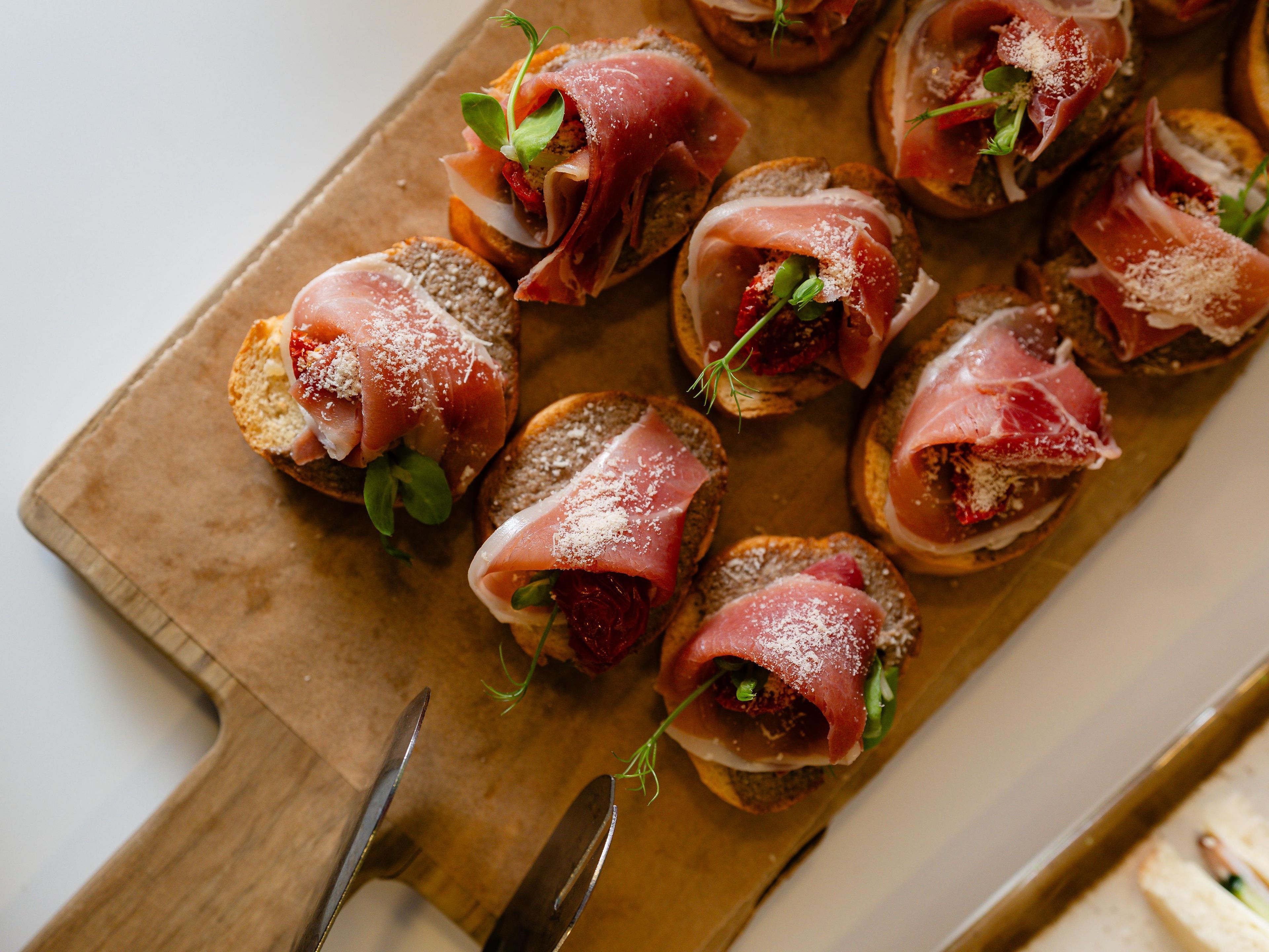 Una tabla de madera con rebanadas de pan cubiertas de prosciutto y vegetales verdes.