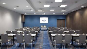 A conference room with rows of tables and chairs, a projector screen, and a carpeted floor.