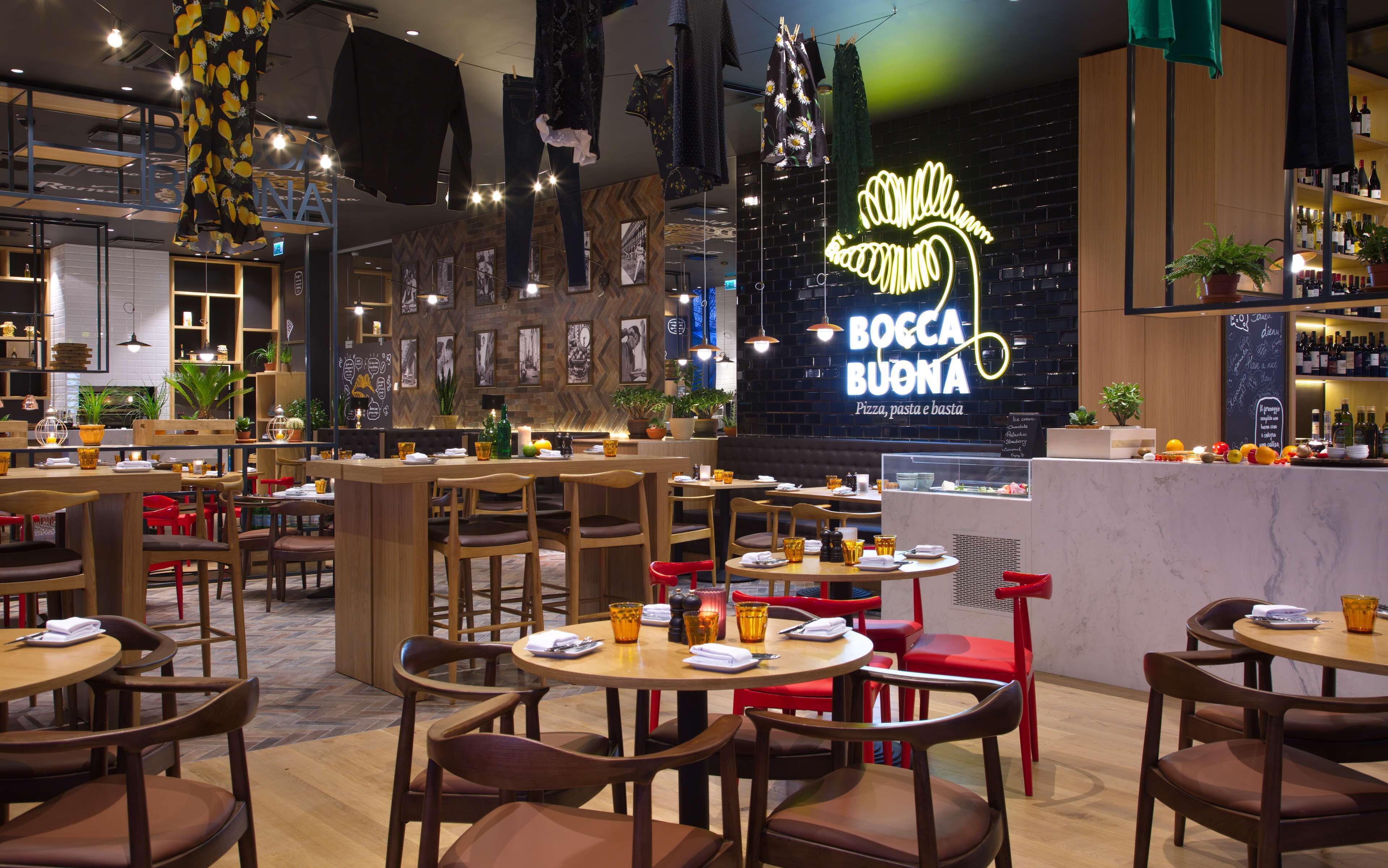 A restaurant interior with multiple dining tables and chairs, a neon sign for 'BOCCA BUONA Pizza, pasta e basta', and a bar area with shelves of bottles.