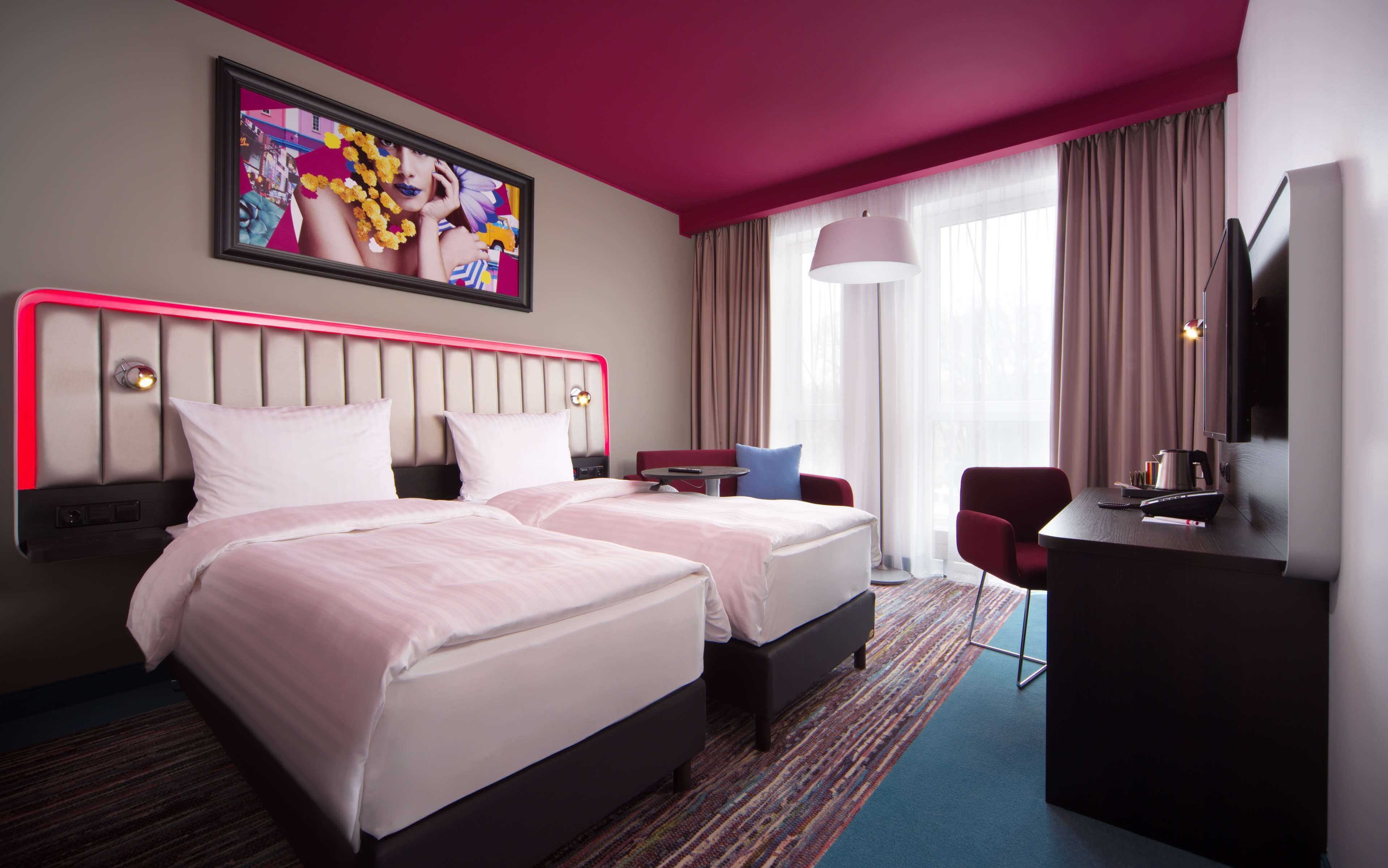 Habitación de hotel con dos camas, un escritorio, una silla, un televisor y un ventanal con cortinas.