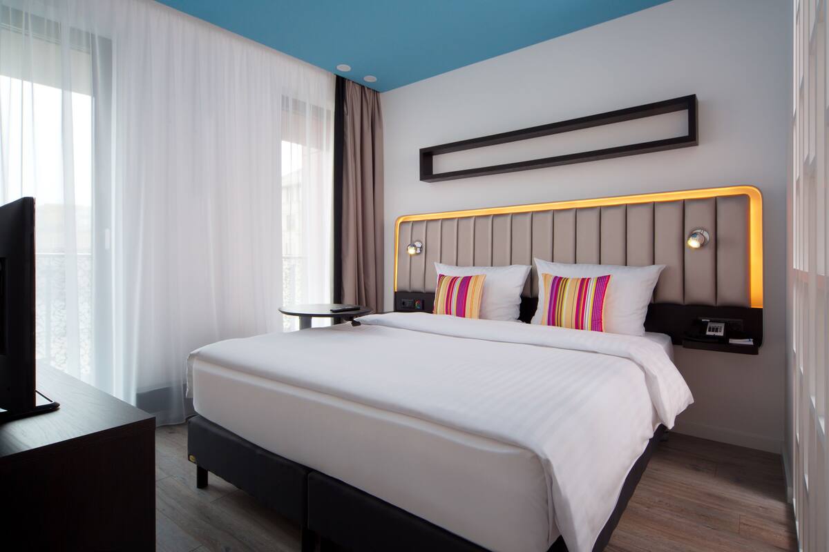 Moderni hotellihuone, jossa on suuri sänky, yöpöytä, televisio ja näkymä ulos.