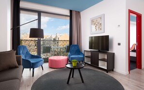 Una sala de estar moderna con un televisor, dos sillas azules, un otoman rosa y una mesita con una planta.
