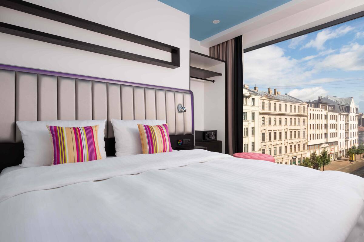 Moderni hotellihuone, jossa on suuri sänky, erikoisella designilla varustettu pääty ja suuri ikkuna, josta avautuu näkymä historiallisiin rakennuksiin.