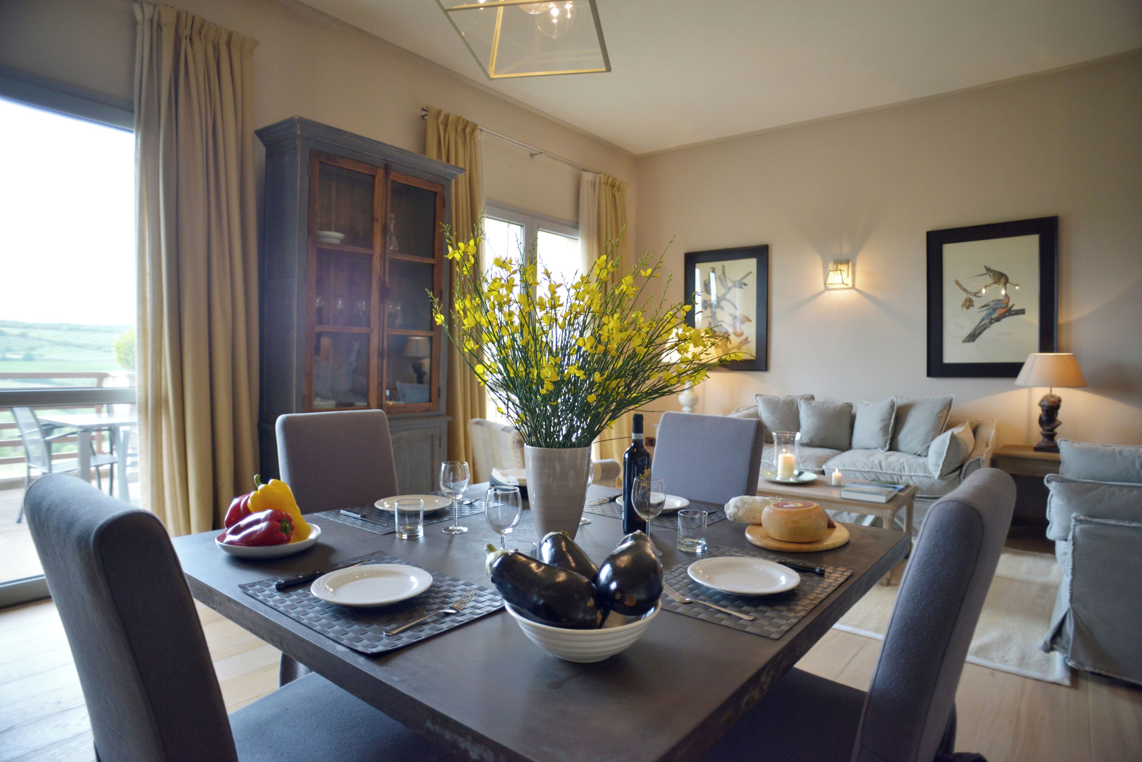 A dining area with a table set for four, a vase with yellow flowers, and a bowl of vegetables.