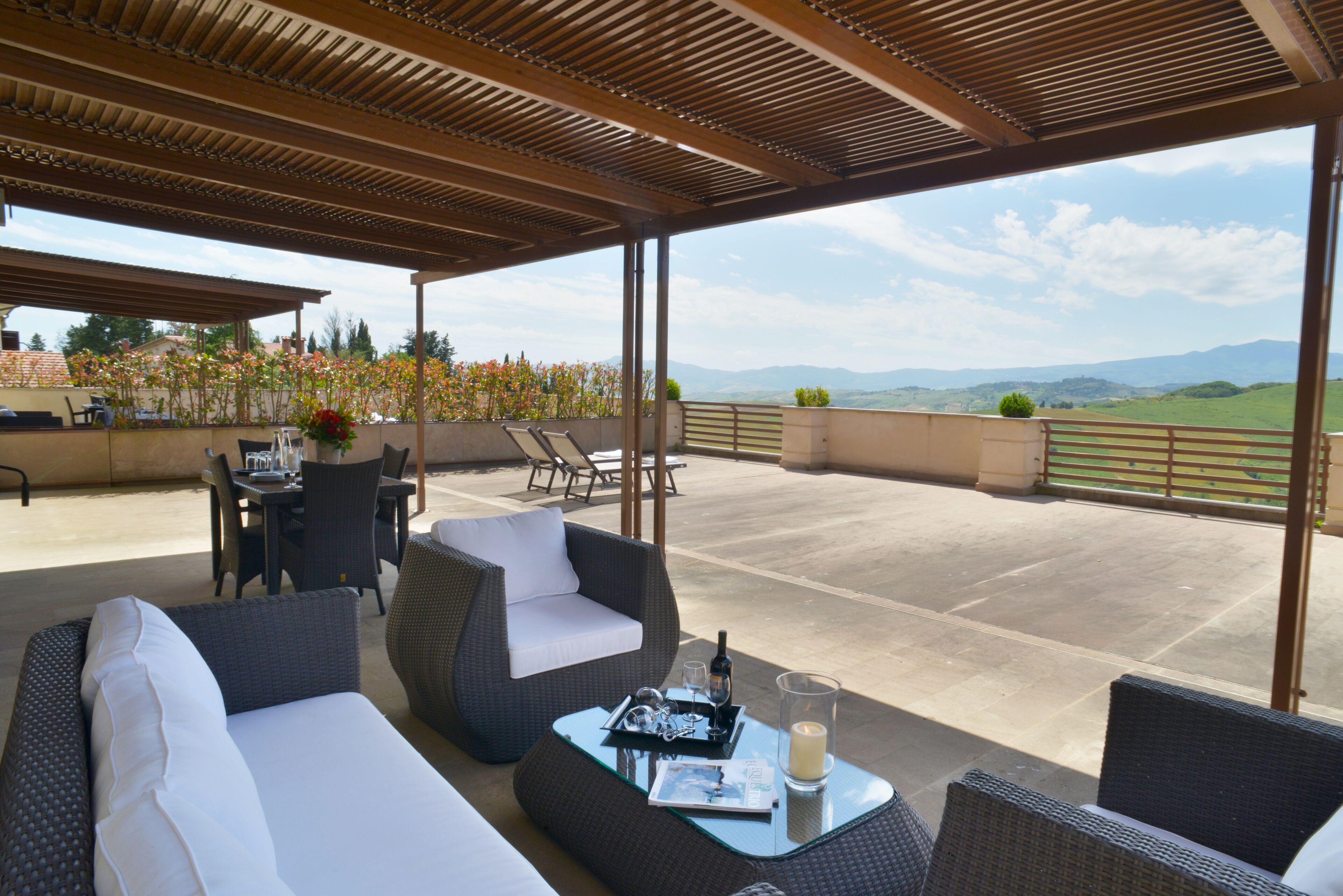 A covered outdoor seating area with wicker furniture, a glass table, and a view of the countryside.