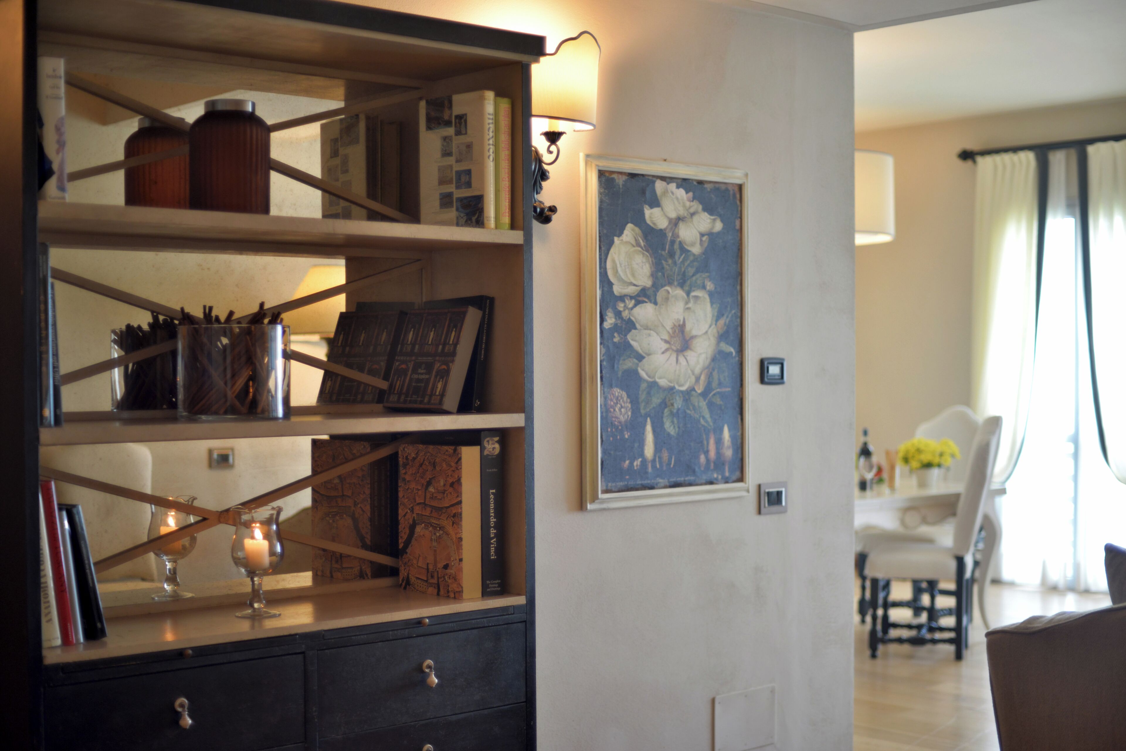 Una habitación con una estantería, un cuadro floral enmarco y un comedor al fondo.
