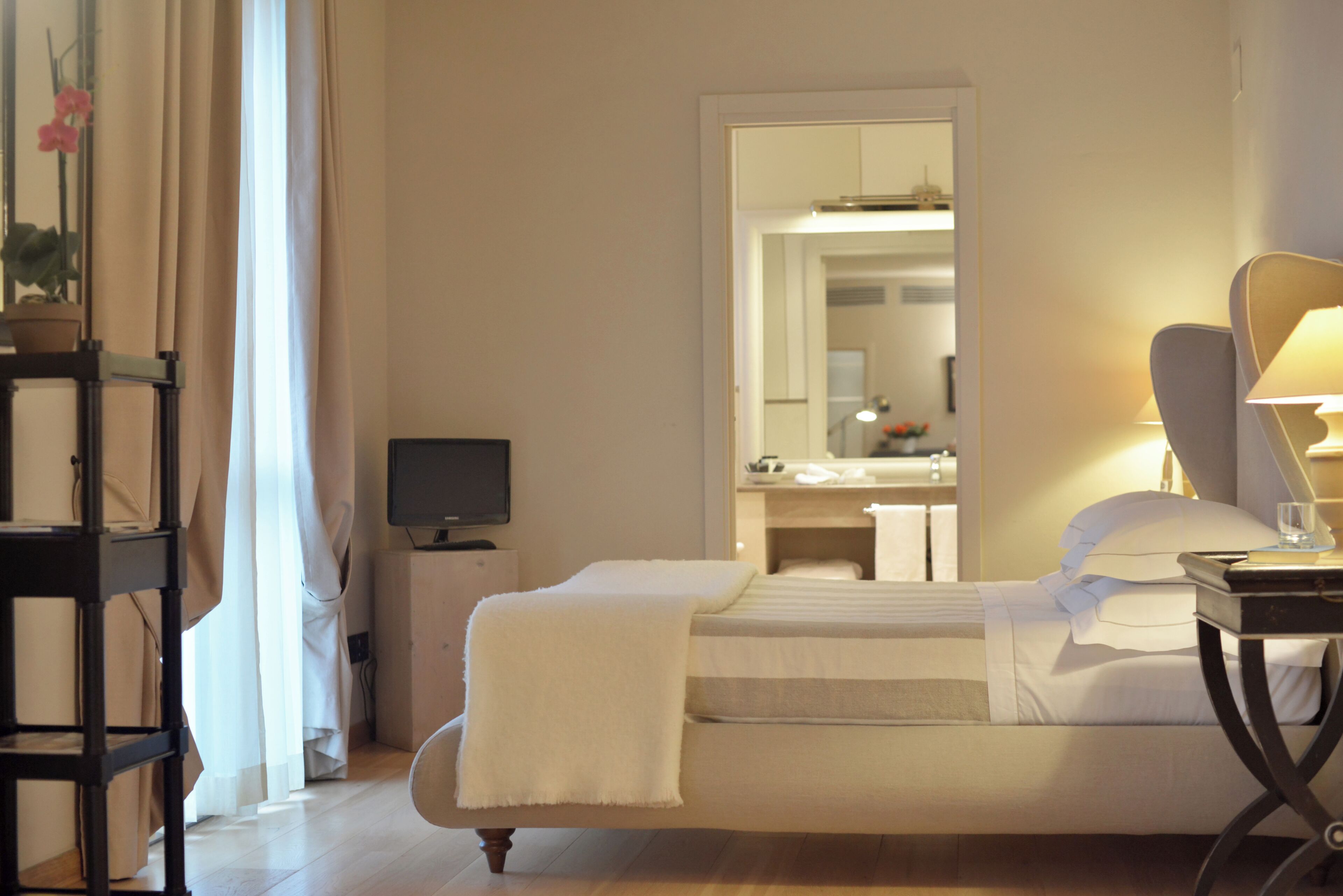 A hotel room with a bed, a desk, a chair, a TV, and a bathroom visible in the mirror reflection.