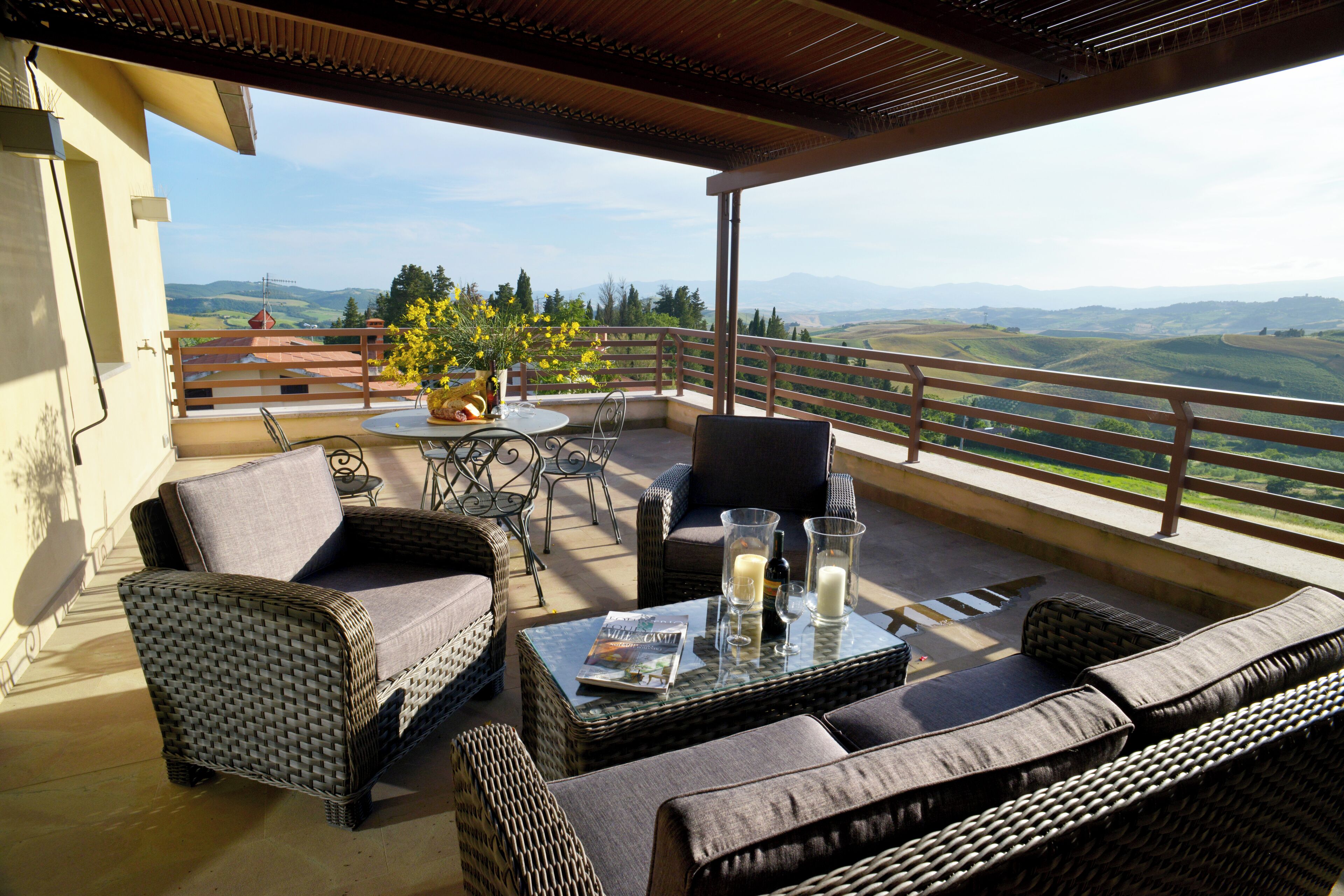 A covered outdoor seating area with wicker furniture, a table with drinks, and a view of rolling hills.