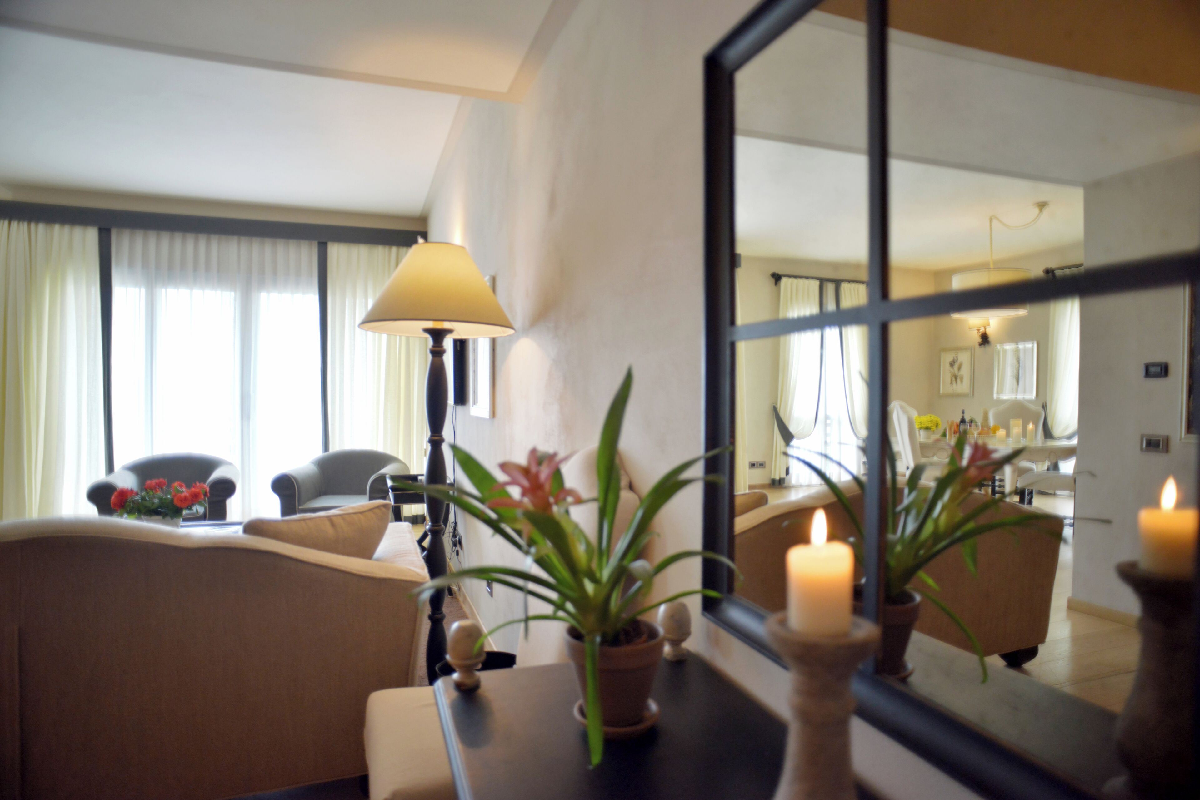 Een woonkamer met een bank, een tafel met een lamp, een potplant en een spiegel die de kamer weerkaatst.