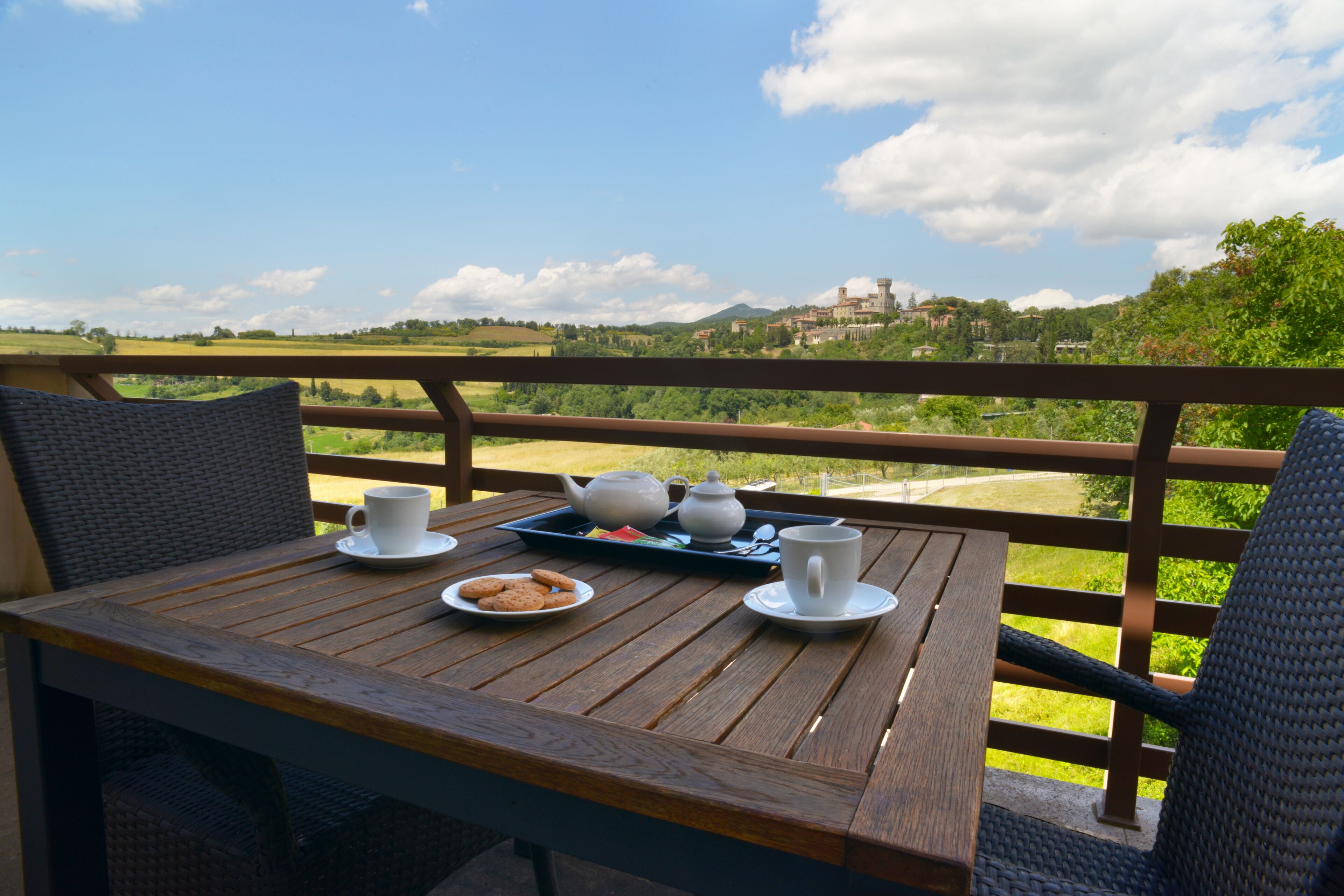 Una mesa de madera con una bandeja de galletas, un calentador de tetera y una taza en un balcón con vistas a un paisaje pintoresco.