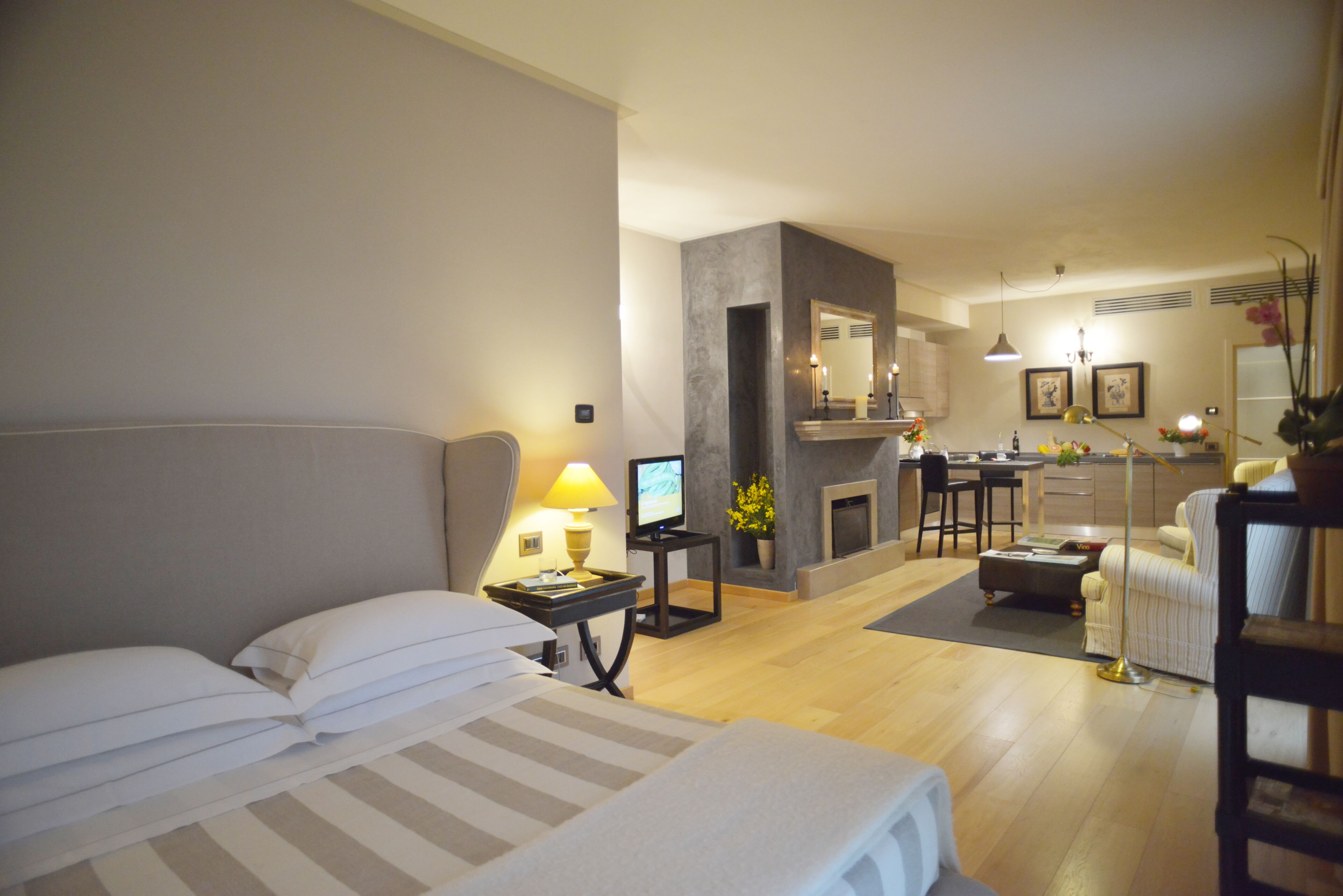 Una camera d'hotel con un letto, comodino, lampada, TV e vista sull'area soggiorno con divano, poltrone e camino.