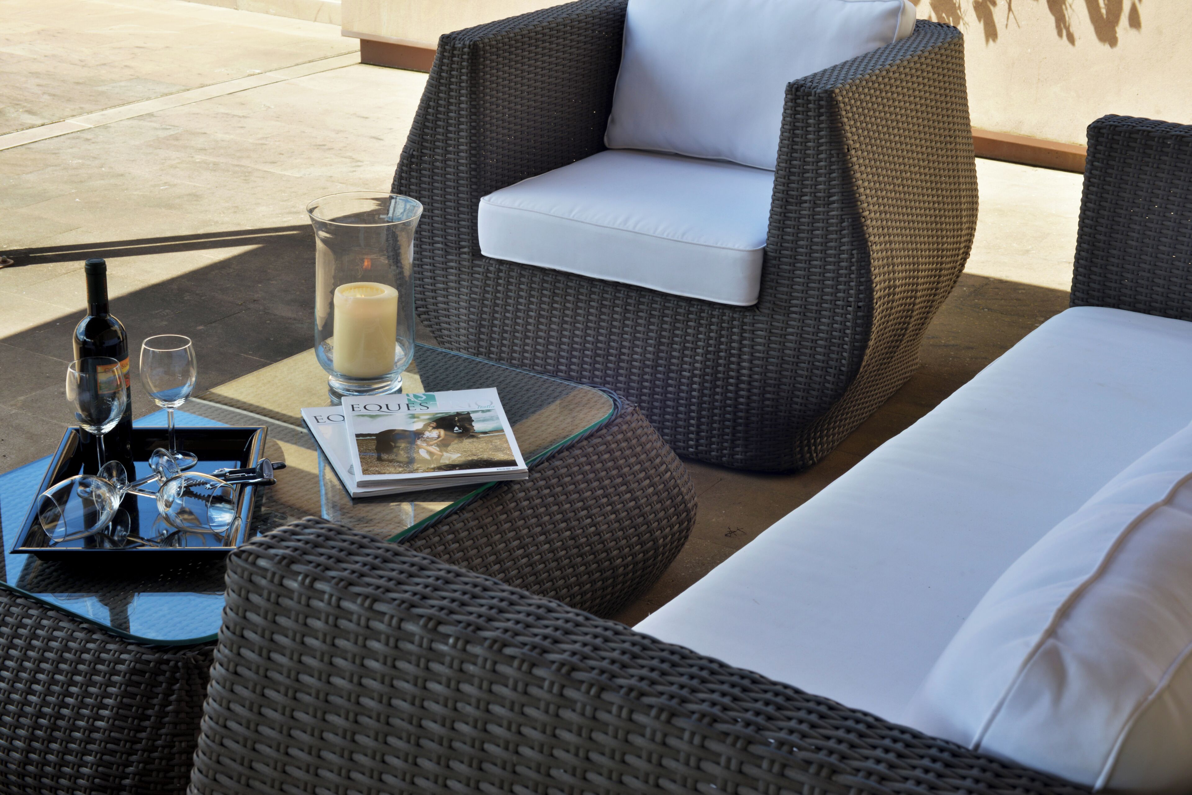 Una silla de mimbre con cojín blanco, una mesa de vidrio con vela, copas de vino y una revista.