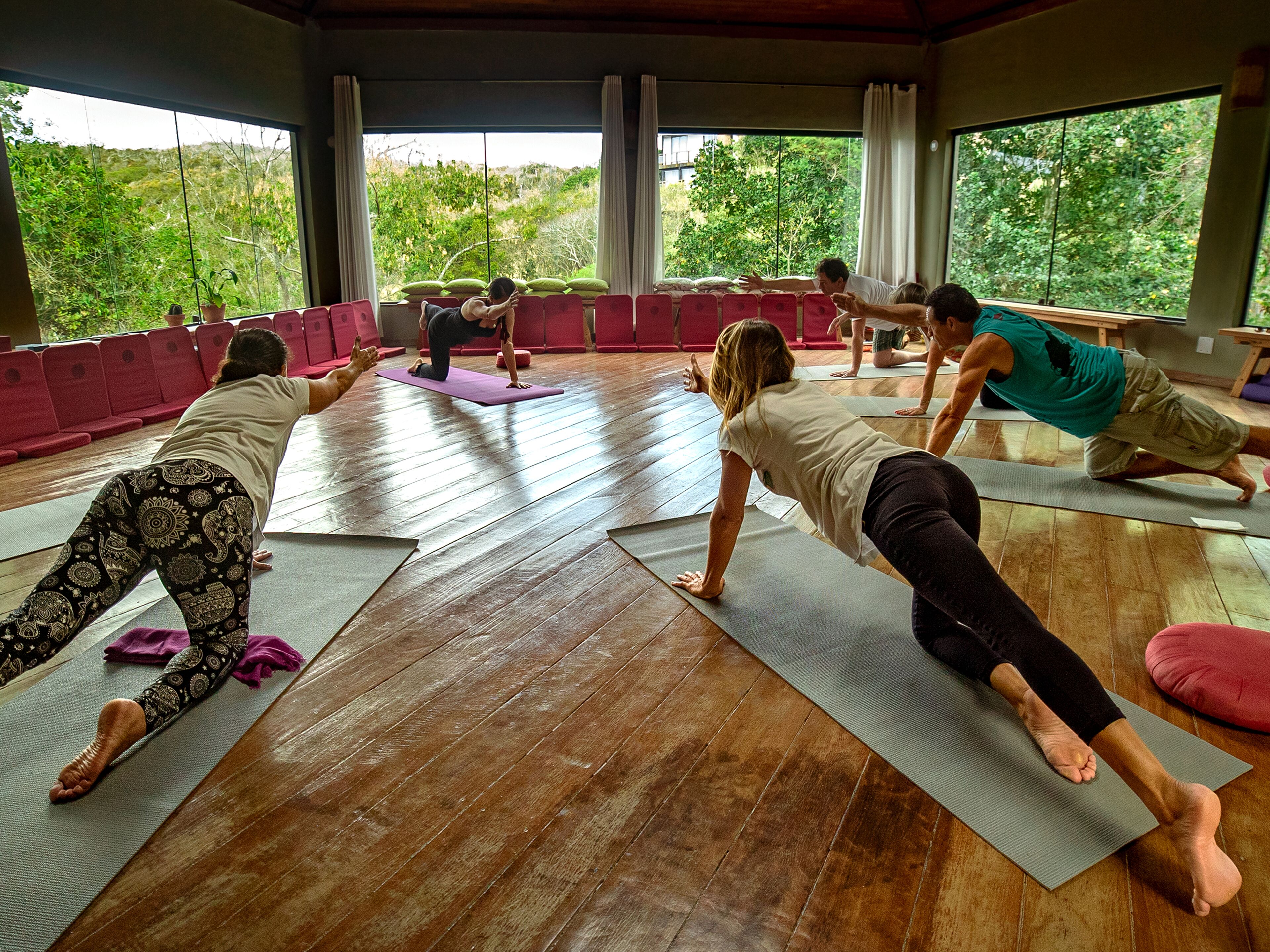 A yoga class in a room with large windows, wooden floors, and red chairs.