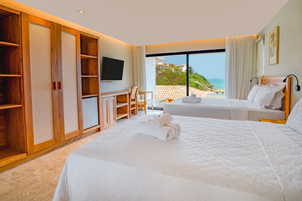 A hotel room with two beds, a desk, and a TV. There is a view of the beach and ocean through large windows.