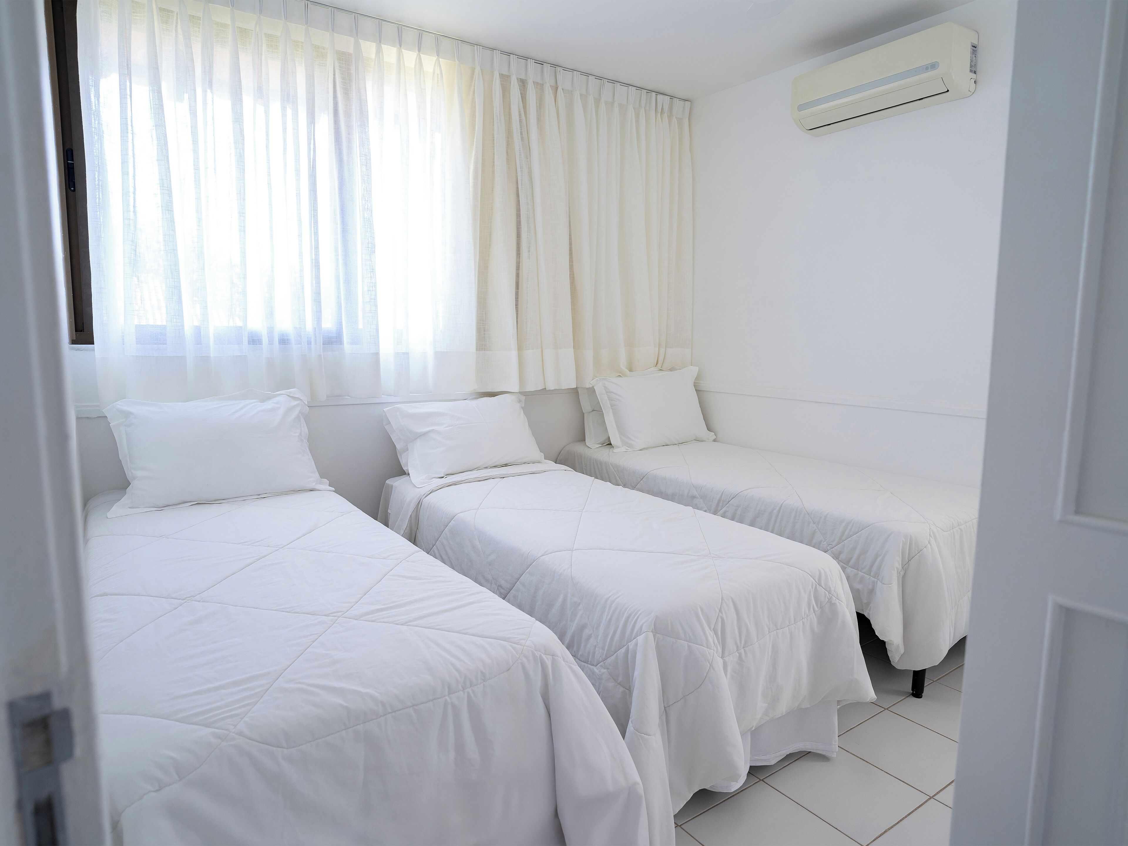 A hotel room with two single beds, a window with sheer curtains, and an air conditioning unit on the wall.