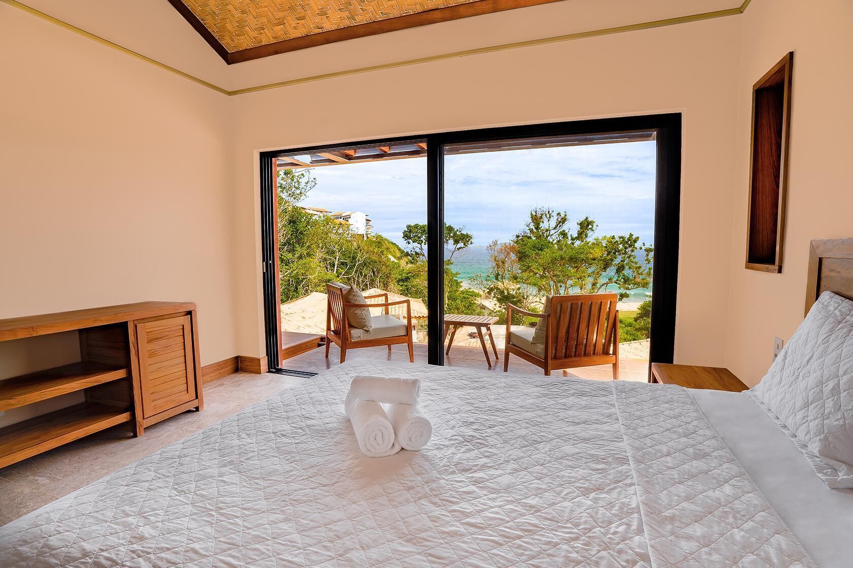 A bedroom with a large bed, a wooden side table, a chair, and a view of the beach through a sliding door.