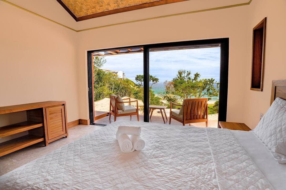 A bedroom with a large bed, a wooden side table, a chair, and a view of the beach through a sliding door.
