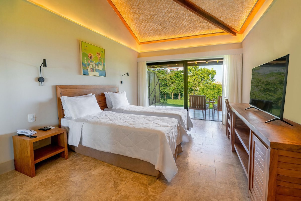 A hotel room with two beds, a TV, a small table, and a balcony with a view of greenery.