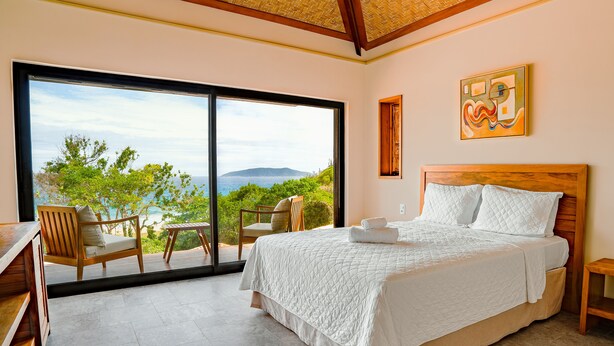 A bedroom with a large bed, a wooden desk, and a chair. There is a view of the ocean and trees through a sliding glass door. - Búzios Espiritualidade Hotel (Búzios)