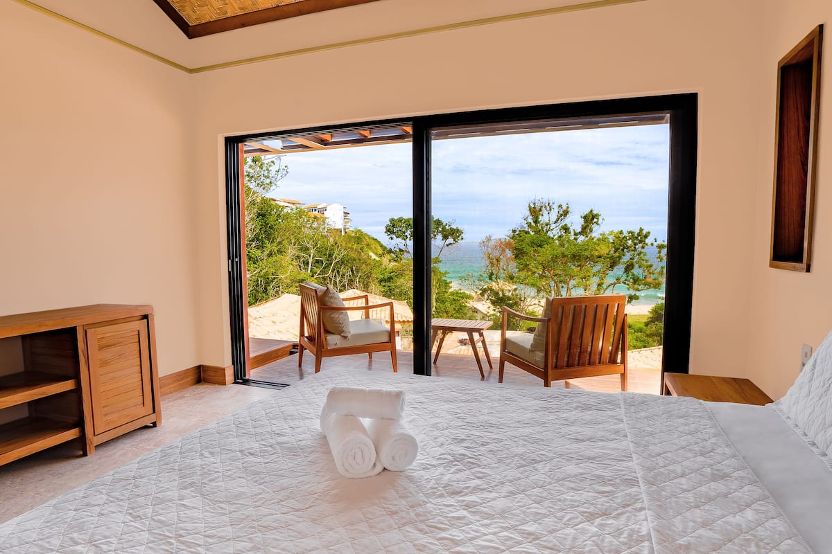 A bedroom with a large bed, a wooden nightstand, a wooden chair, and a view of the beach and ocean through a sliding door.