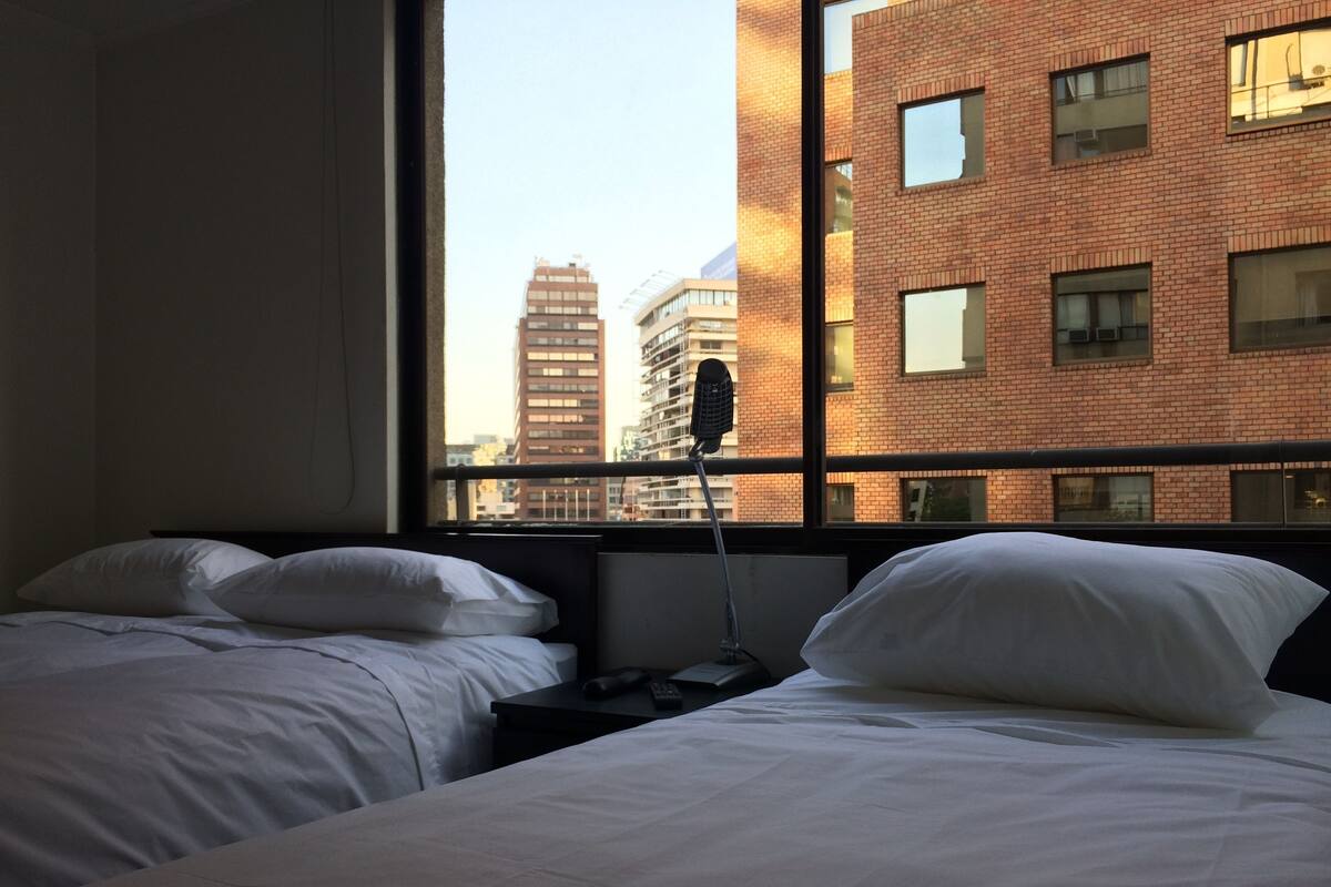 Habitación de hotel con dos camas, una ventana que ofrece vistas a un edificio de ladrillo y una mesita de noche con lámpara.