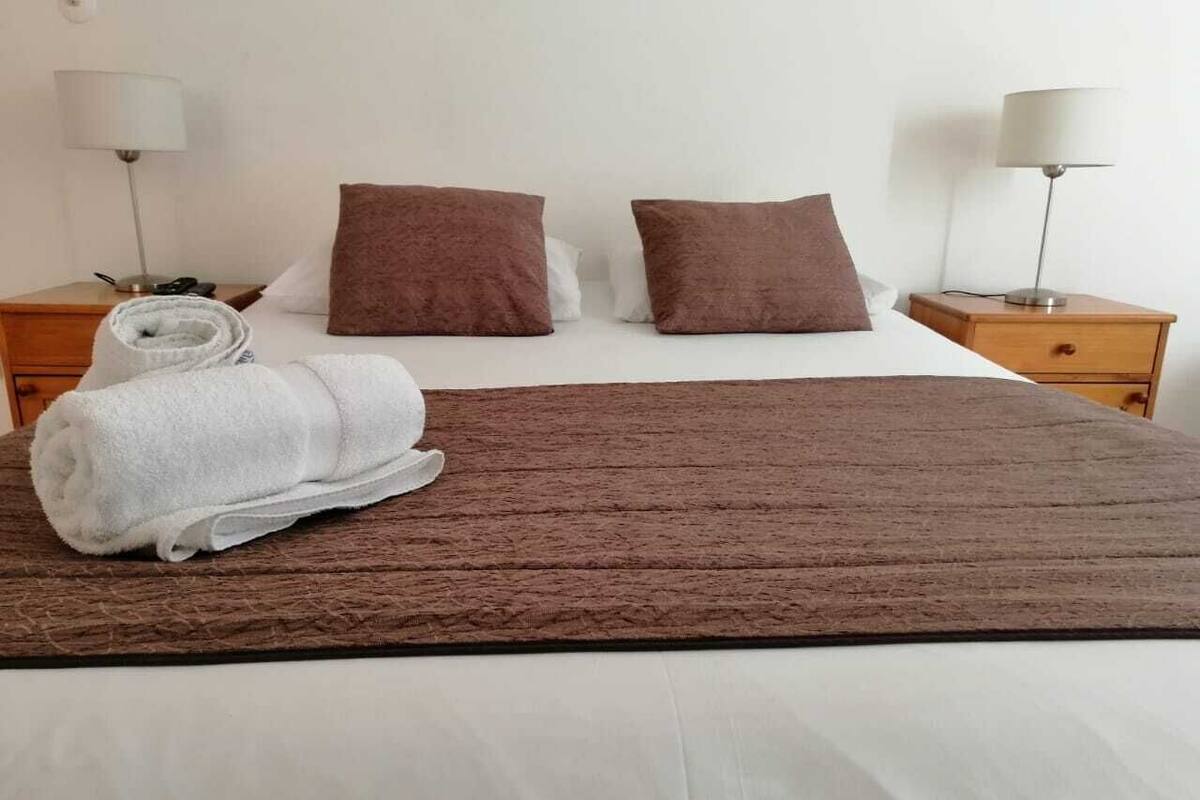 Una cama bien hecha con una colcha marrón, dos almohadas y una toalla blanca enrollada al lado. Hay dos mesitas de noche con lámparas y un mueble auxiliar con una lámpara.