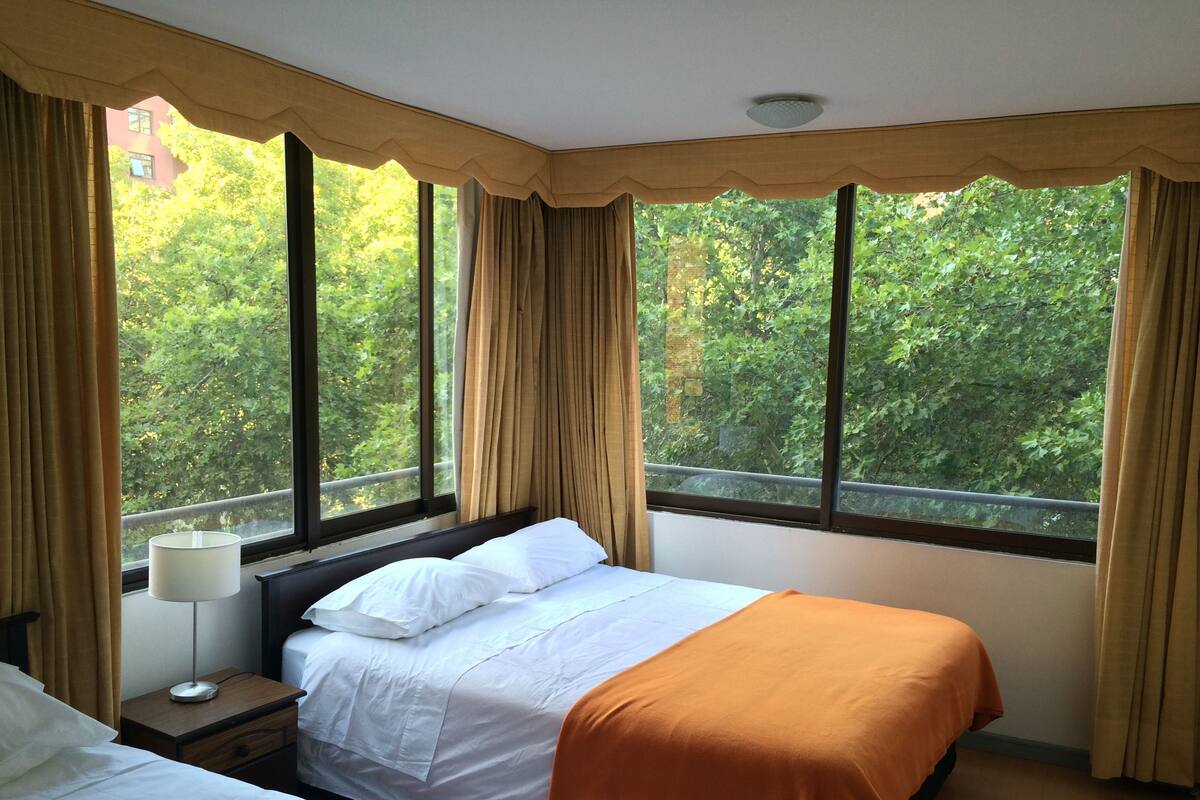 Una habitación de hotel con cama, mesita de noche, lámpara y ventanas amplias con cortinas.