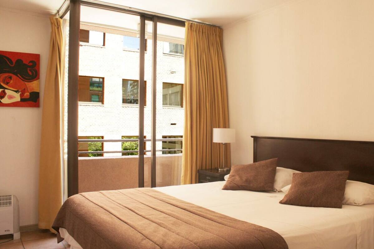 Habitación de hotel con cama, mesita de noche, lámpara, ventana con cortinas y un cuadro en la pared.