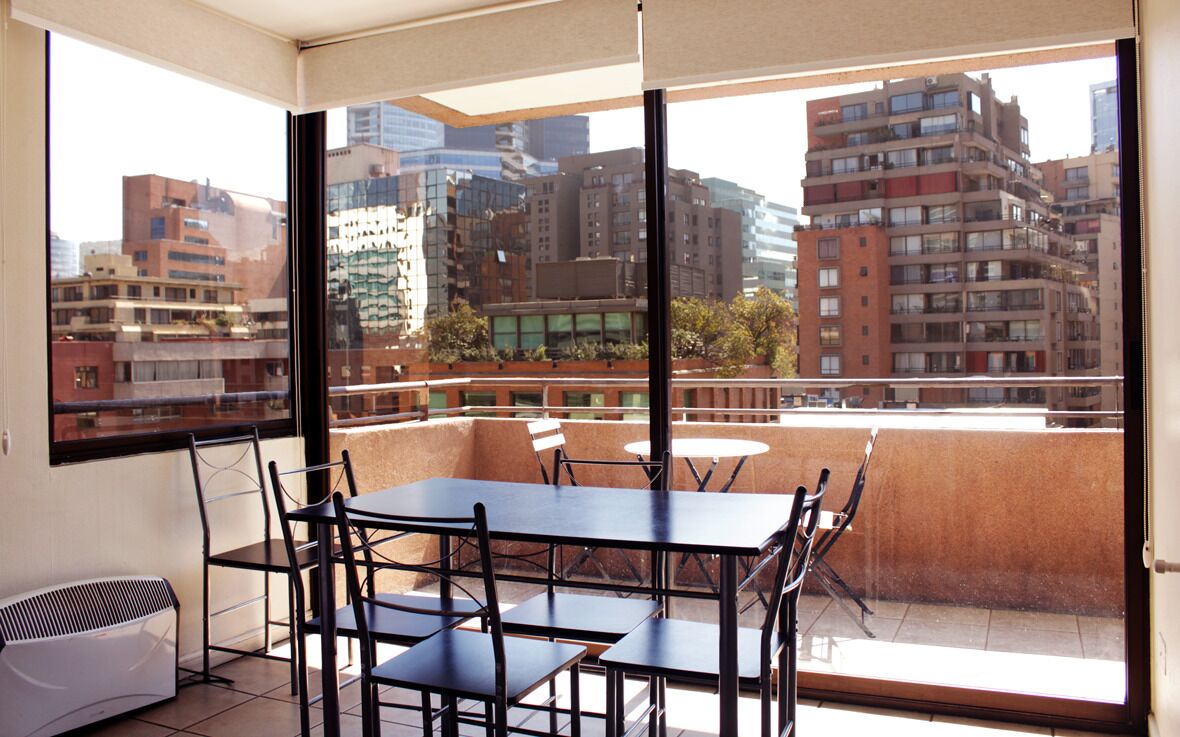 A balcony with a table and chairs, large windows offering a view of city buildings.