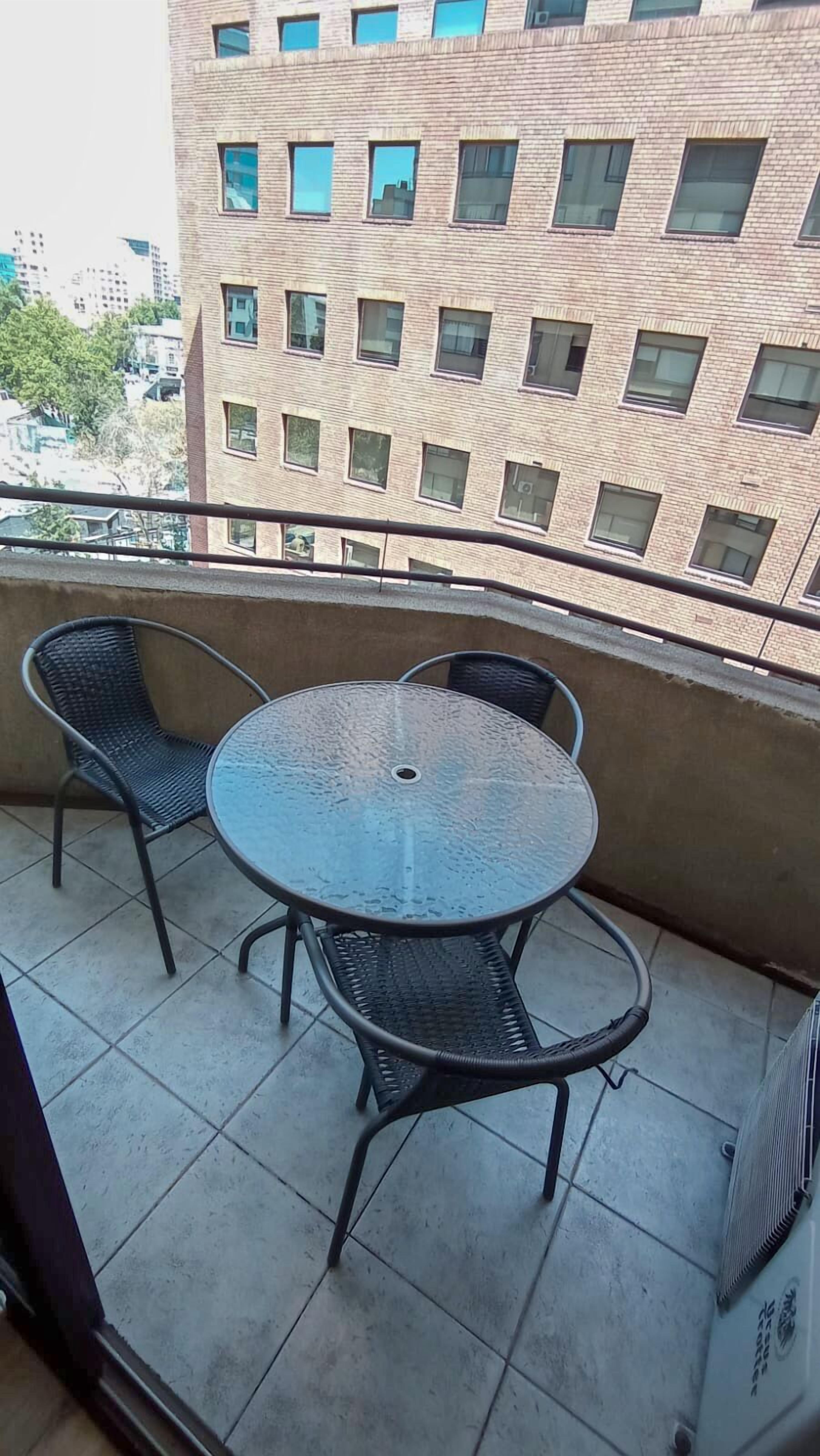 A balcony with a round table and two chairs, a tiled floor, and a view of a brick building with multiple windows.