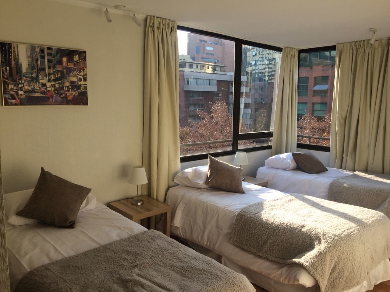 A hotel room with three beds, a painting on the wall, and a view of buildings through large windows.