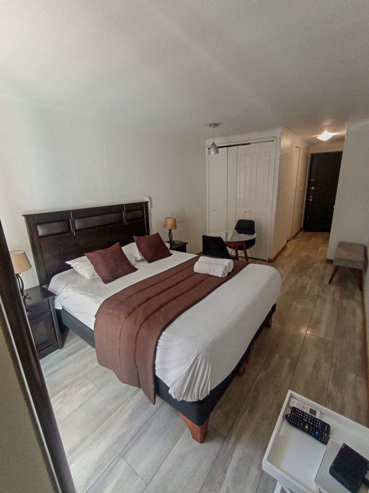 A neatly made bed with a brown blanket, a bedside table with a lamp, and a small table with a remote control.