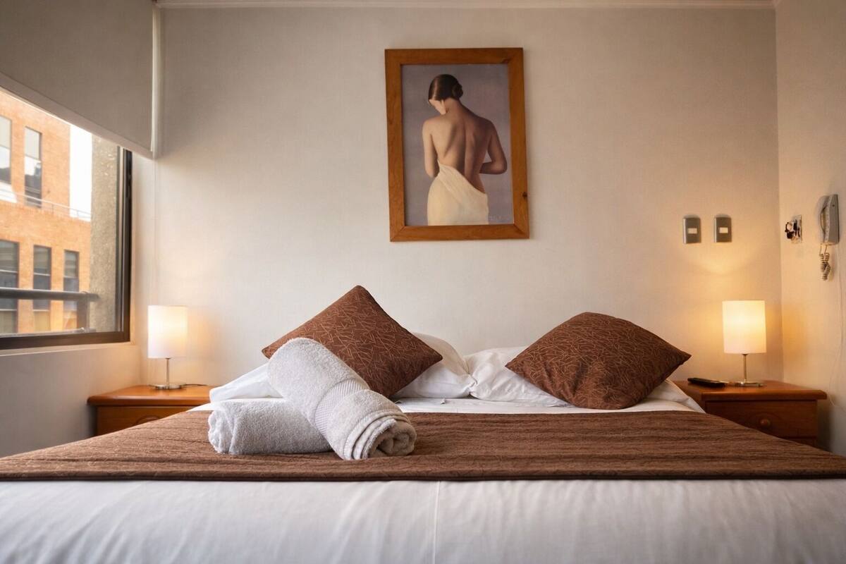 Una habitación de hotel con una cama grande, dos mesitas de noche, una ventana y un cuadro en la pared.
