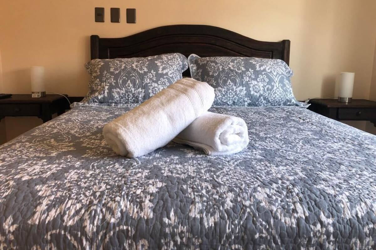 Una cama bien hecha con un edredón estampado de flores, dos almohadas, una mesita de noche con una lámpara y un aplique en la pared.