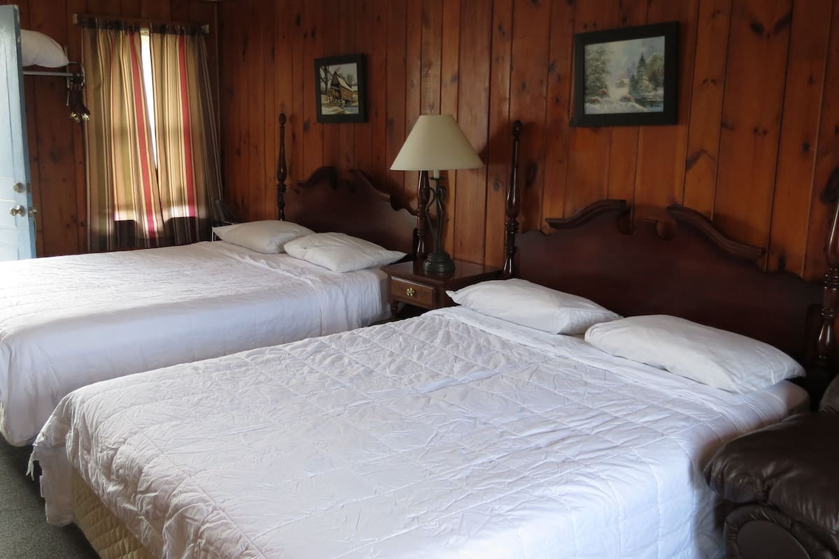 A room with two beds, wooden walls, a window with curtains, and a clock on the wall.