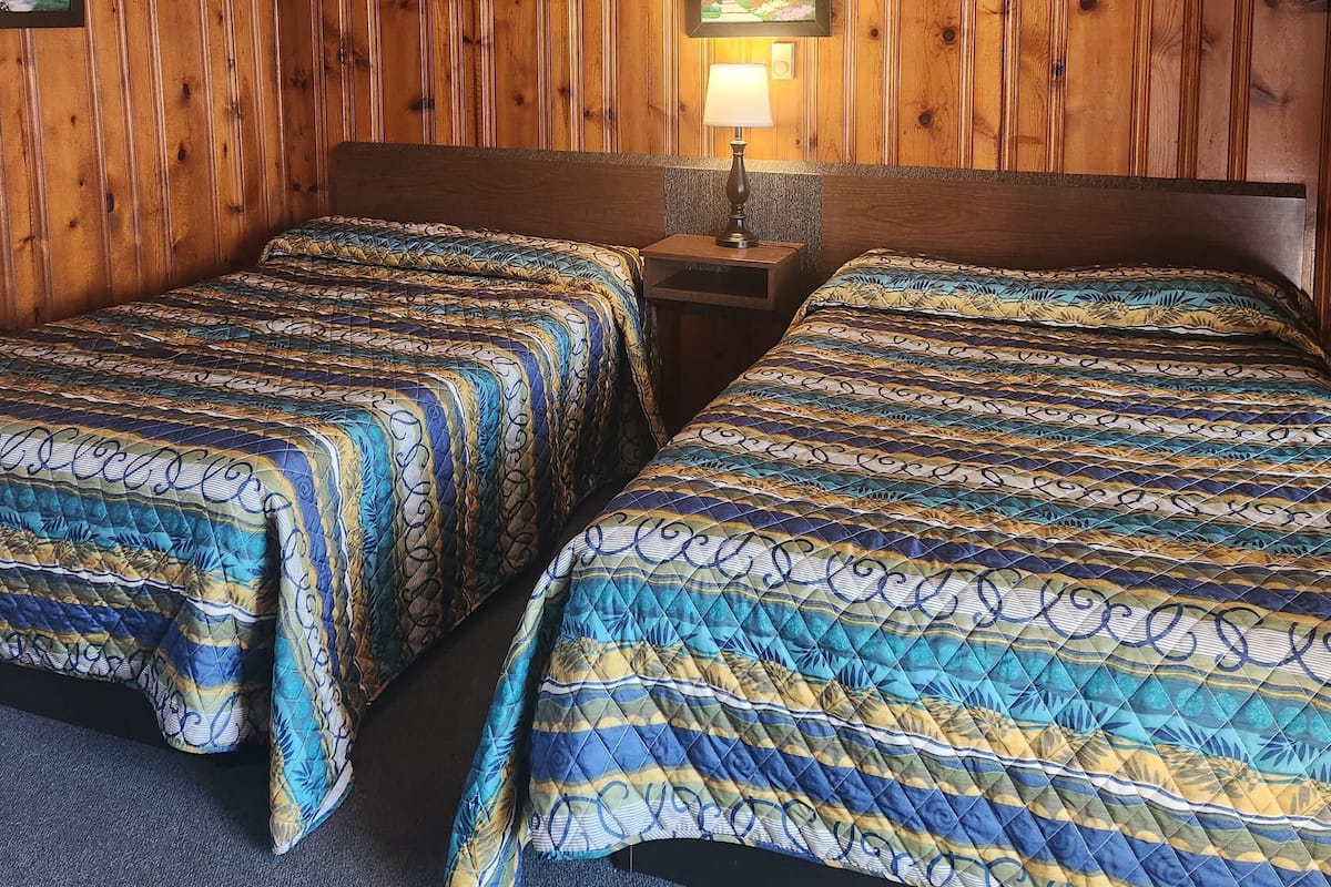 A room with two beds, wooden paneling, a picture on the wall, and a nightstand with a lamp.