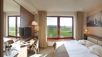 A hotel room with a large bed, a desk with a TV, a view of a green landscape through the windows, and a painting on the wall.